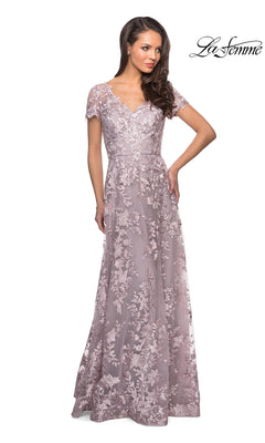 A woman poses with one hand on her hip, modeling the La Femme 27870—a floor-length, short-sleeved lavender lace mother of the bride dress adorned with floral patterns. The “La Femme” logo appears in the top right corner.
