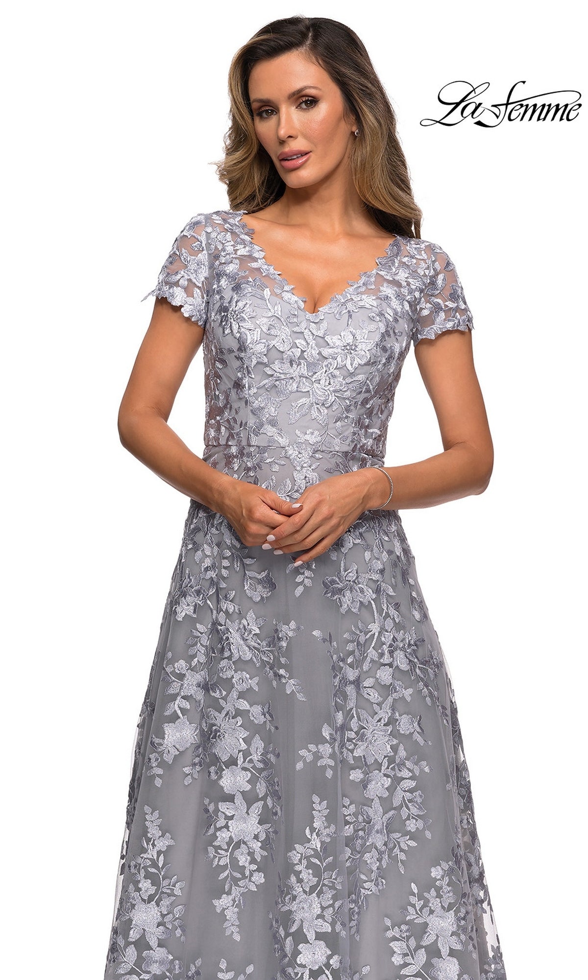 A woman wearing the La Femme 27870 long formal dress—grey, short-sleeved, with a V-neck and floral lace embroidery—stands against a white background. The “La Femme” logo appears in the top right corner.