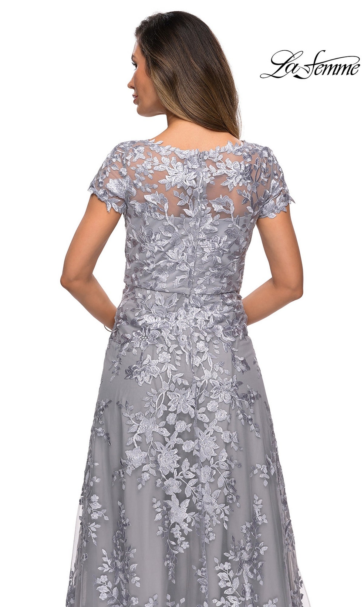 A woman wearing the Long Formal Dress: La Femme 27870—a gray lace mother of the bride gown featuring floral embroidery, short sleeves, and a sheer back detail—stands with her back to the camera. The La Femme logo appears in the top right corner.