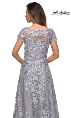 A woman wearing the Long Formal Dress: La Femme 27870—a gray lace mother of the bride gown featuring floral embroidery, short sleeves, and a sheer back detail—stands with her back to the camera. The La Femme logo appears in the top right corner.