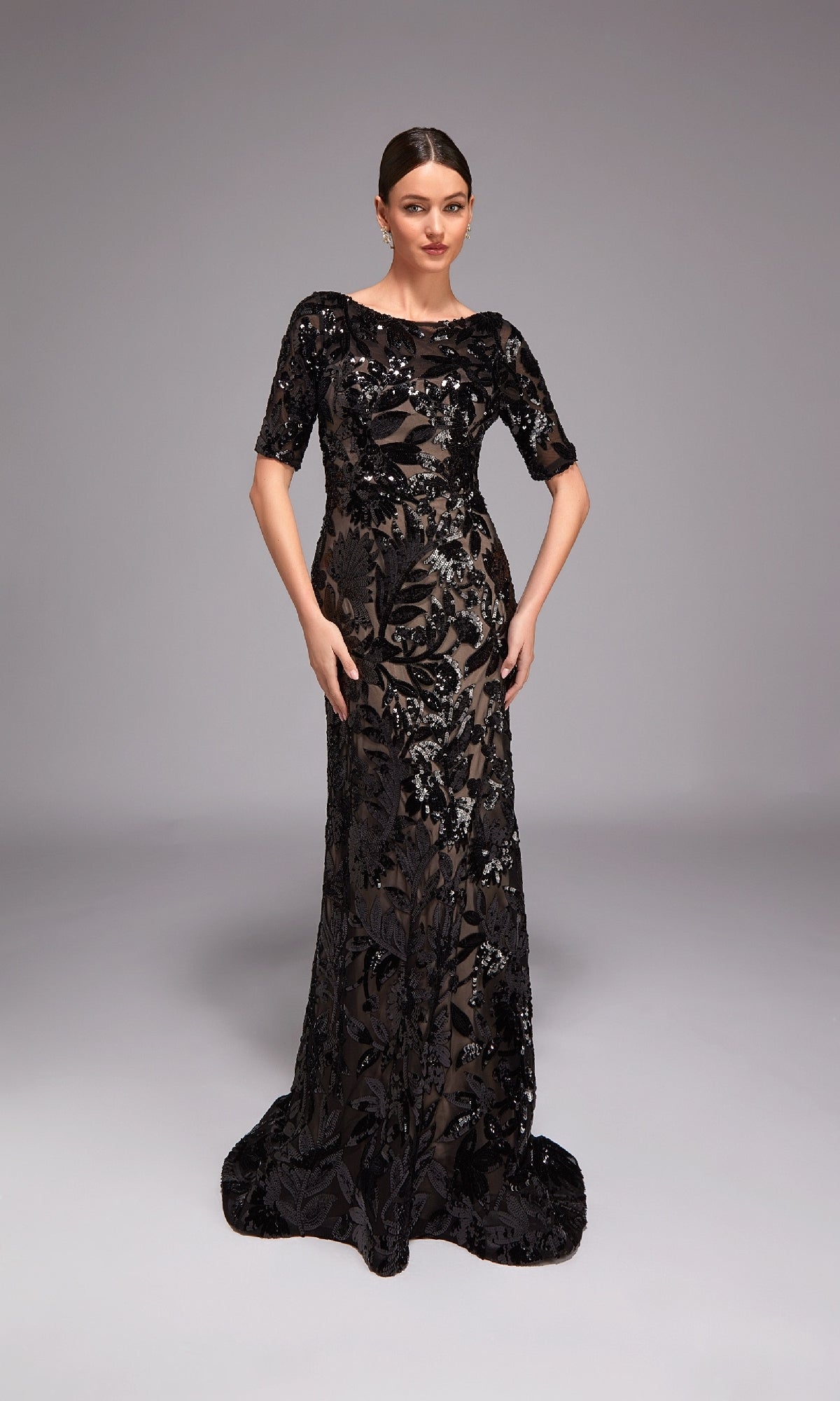 A woman poses against a plain gray background wearing the Alyce 27876 long formal dress—a floor-length black gown with short sleeves, sheer accents, and sequin floral detailing.