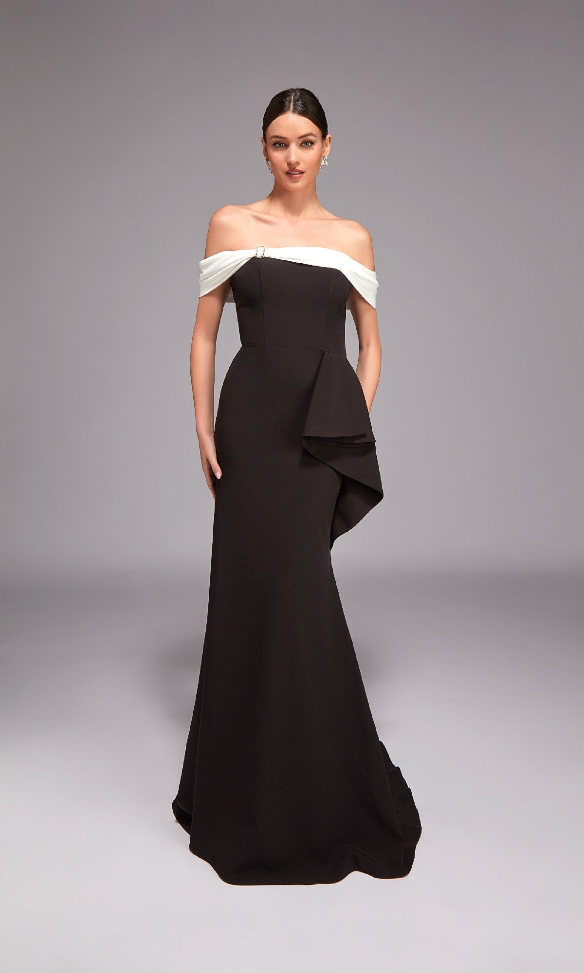 A woman stands in a studio wearing the Alyce 27877 long formal dress, with an off-the-shoulder white folded neckline, asymmetric peplum detail, and a sleek black mermaid silhouette.