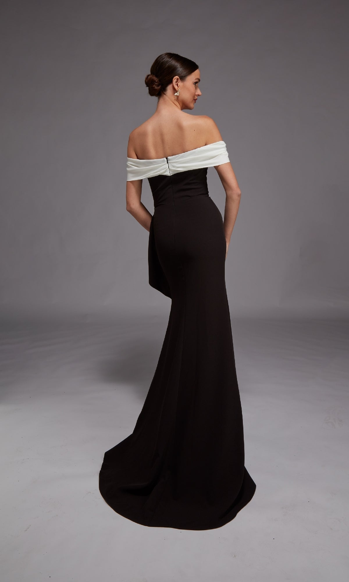 A woman stands with her back to the camera, wearing the Alyce 27877 long black formal dress featuring a white fold-over neckline and elegant mermaid silhouette, set against a plain gray backdrop.