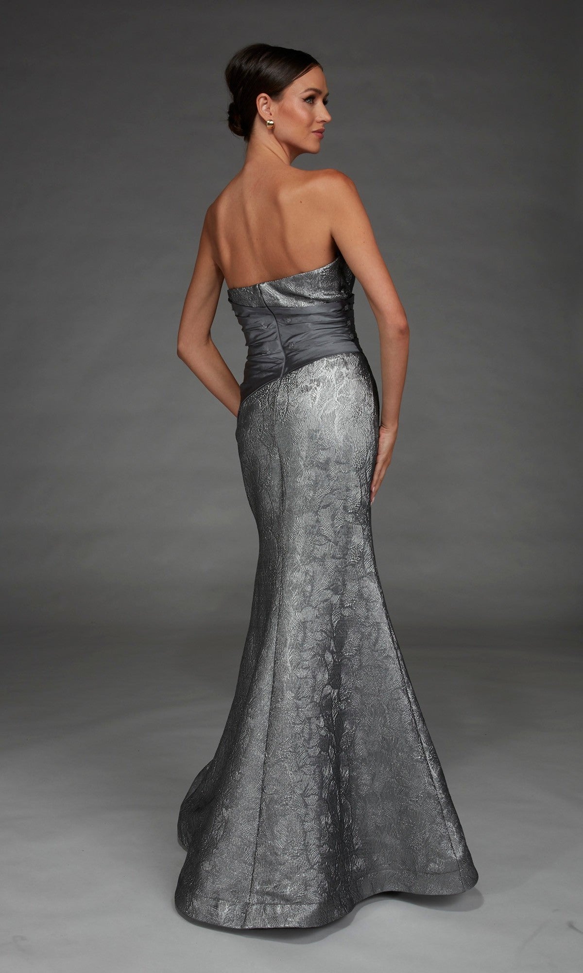 A woman stands facing away against a gray background, wearing the Alyce 27881 long formal dress—a strapless metallic silver mermaid evening gown with a fitted bodice and flared skirt.