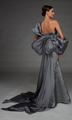 Woman wearing the Alyce 27881 long formal dress, crafted in silver jacquard with a strapless neckline, dramatic oversized bow accent, and an elegant long train, posing against a plain gray background.