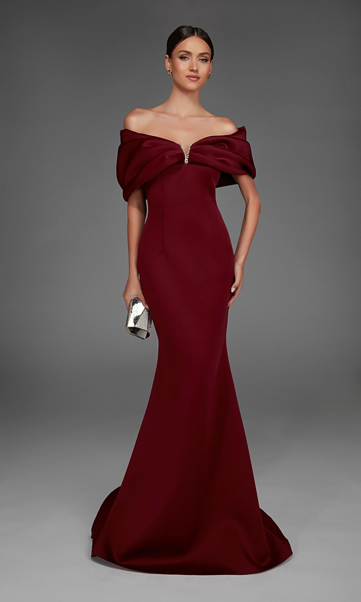 A woman wearing the Alyce 27883 Long Formal Dress in burgundy, featuring an off-the-shoulder neckline and a graceful floor-length mermaid skirt, holds a silver clutch while standing against a plain gray background.