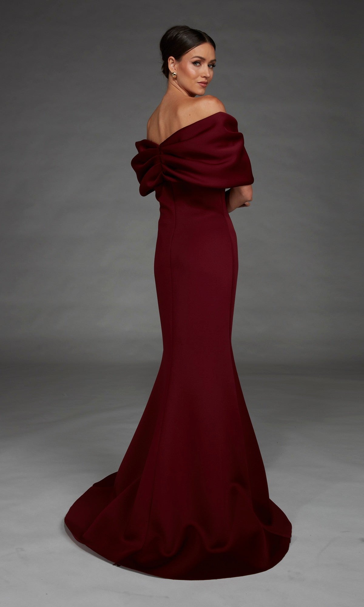 A woman in the Alyce 27883 long formal dress, an elegant off-the-shoulder burgundy gown with a striking large bow detail at the back and a flowing mermaid skirt, stands against a plain grey background.