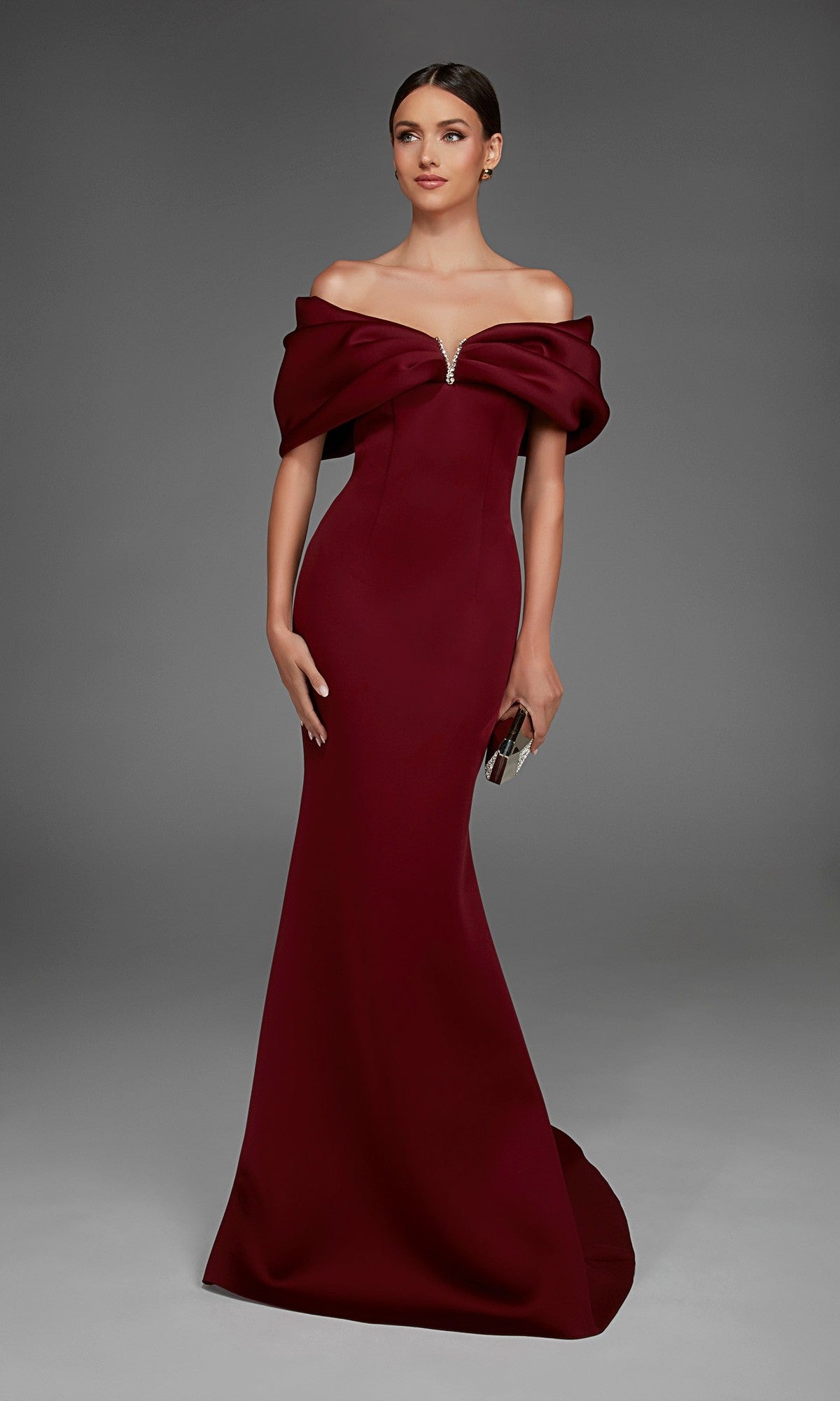 A woman wearing the Alyce 27883 long formal dress—a floor-length, off-the-shoulder burgundy gown with a graceful mermaid skirt—holds a small silver clutch while posing against a plain gray background.
