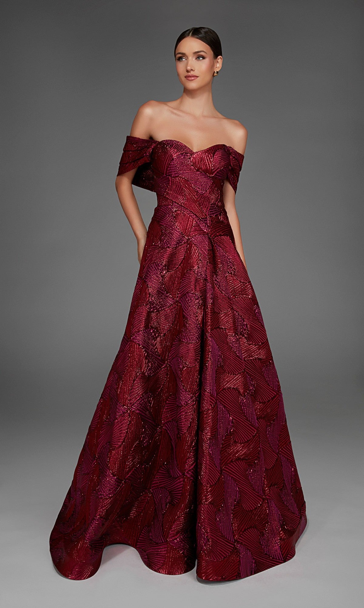A woman poses against a plain gray background wearing the Alyce 27884, a long formal dress featuring an off-the-shoulder neckline, elegant jacquard fabric, and an A-line skirt.