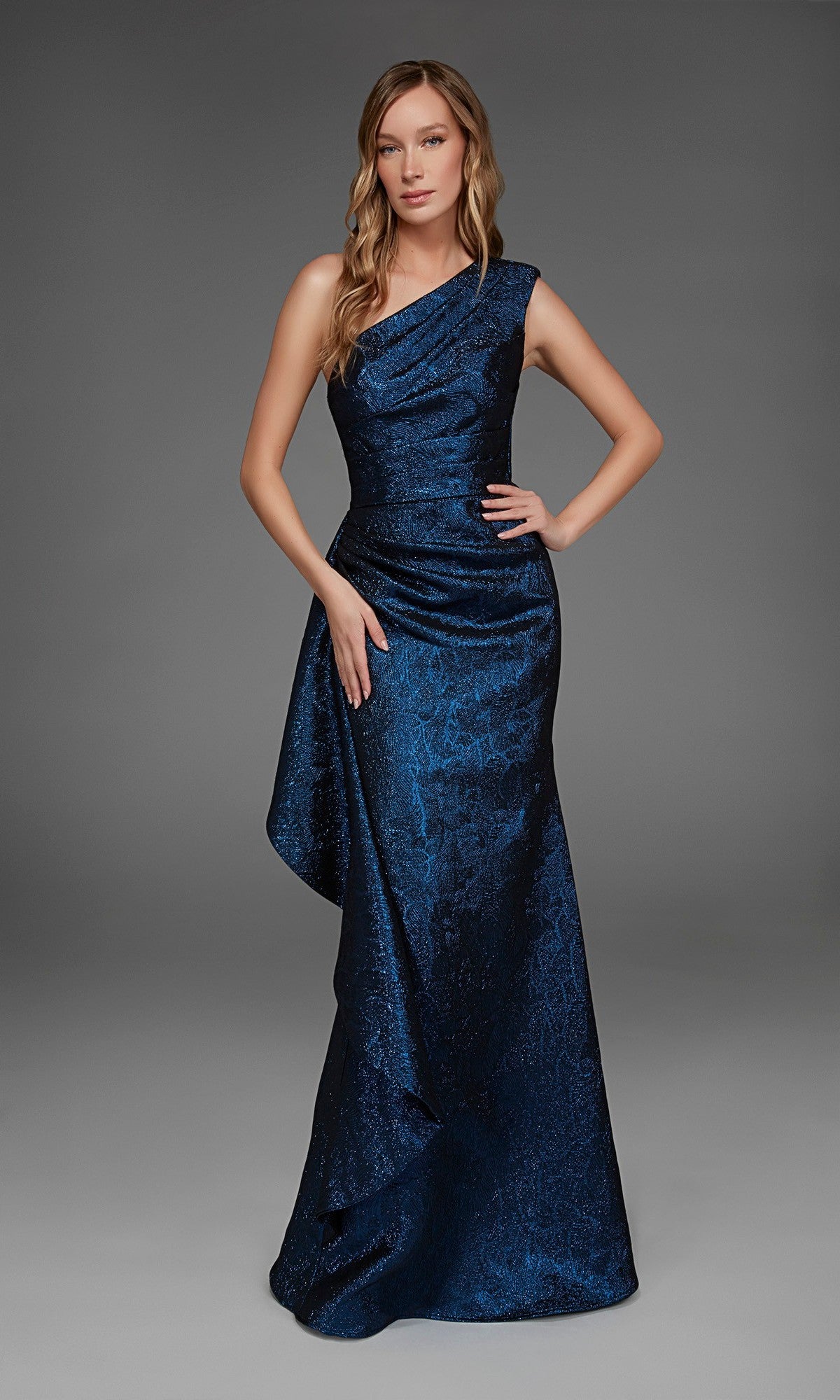 A woman stands against a plain gray background wearing the Alyce 27885, a long formal dress featuring a floor-length, one-shoulder design in dark blue with a textured pattern.