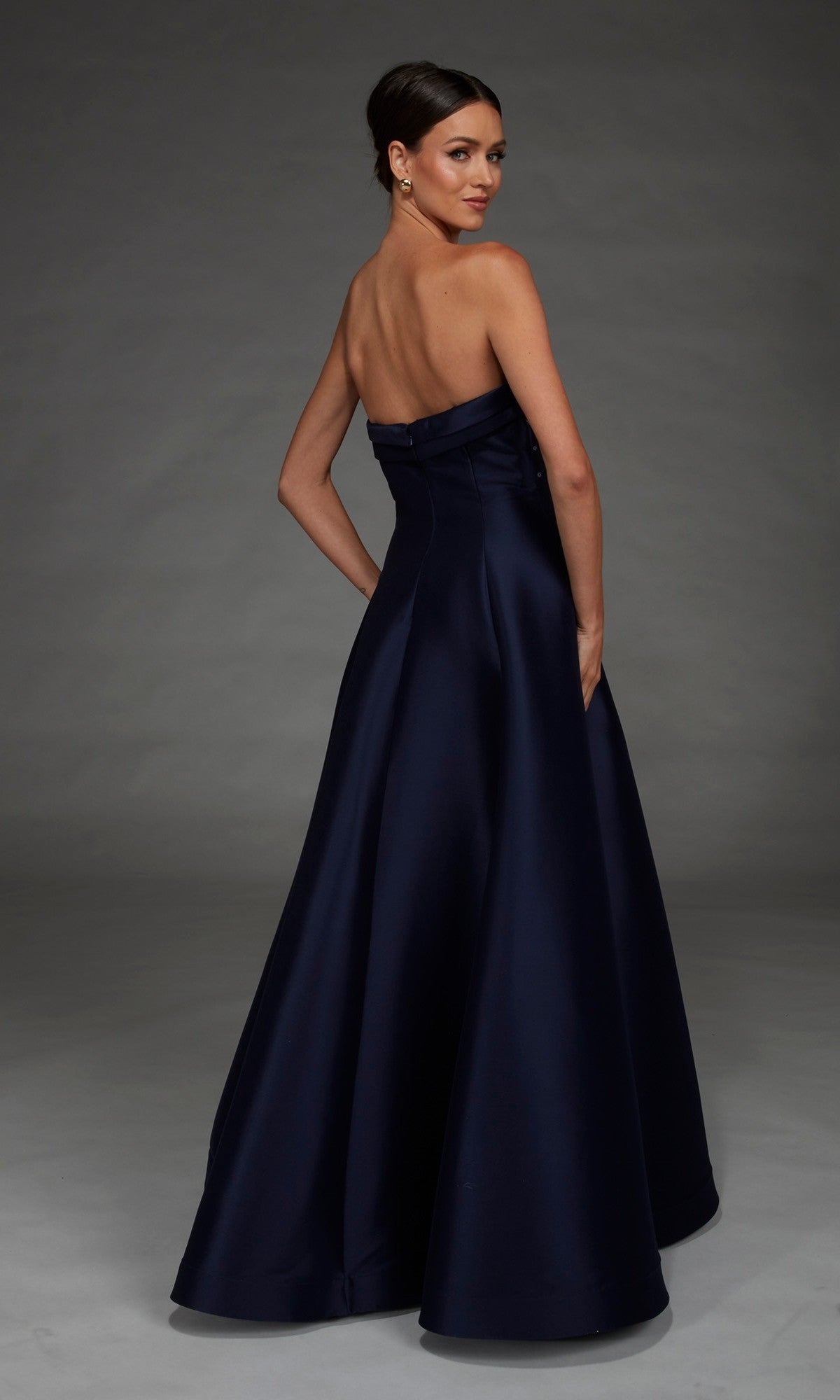 Wearing the Alyce 27887 Long Formal Dress with a strapless neckline and floor-length navy blue silhouette, the woman stands facing away and glances over her shoulder against a plain gray background.
