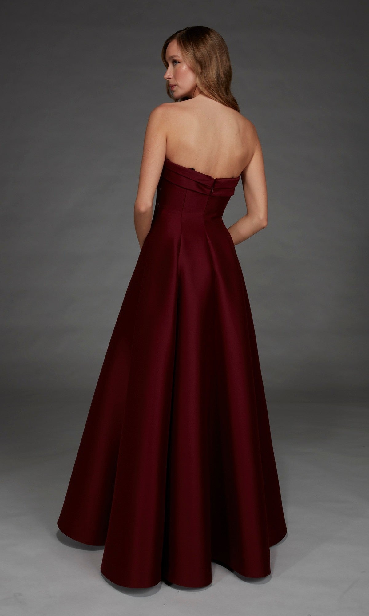 A woman stands with her back turned against a gray studio backdrop, wearing the Alyce 27887 long formal dress—floor-length, strapless, and dark red.