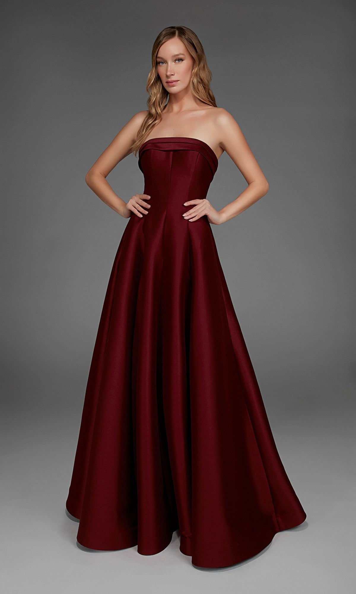 A woman showcases the Long Formal Dress: Alyce 27887, a strapless, floor-length burgundy evening gown, posing with hands on hips against a plain gray background.