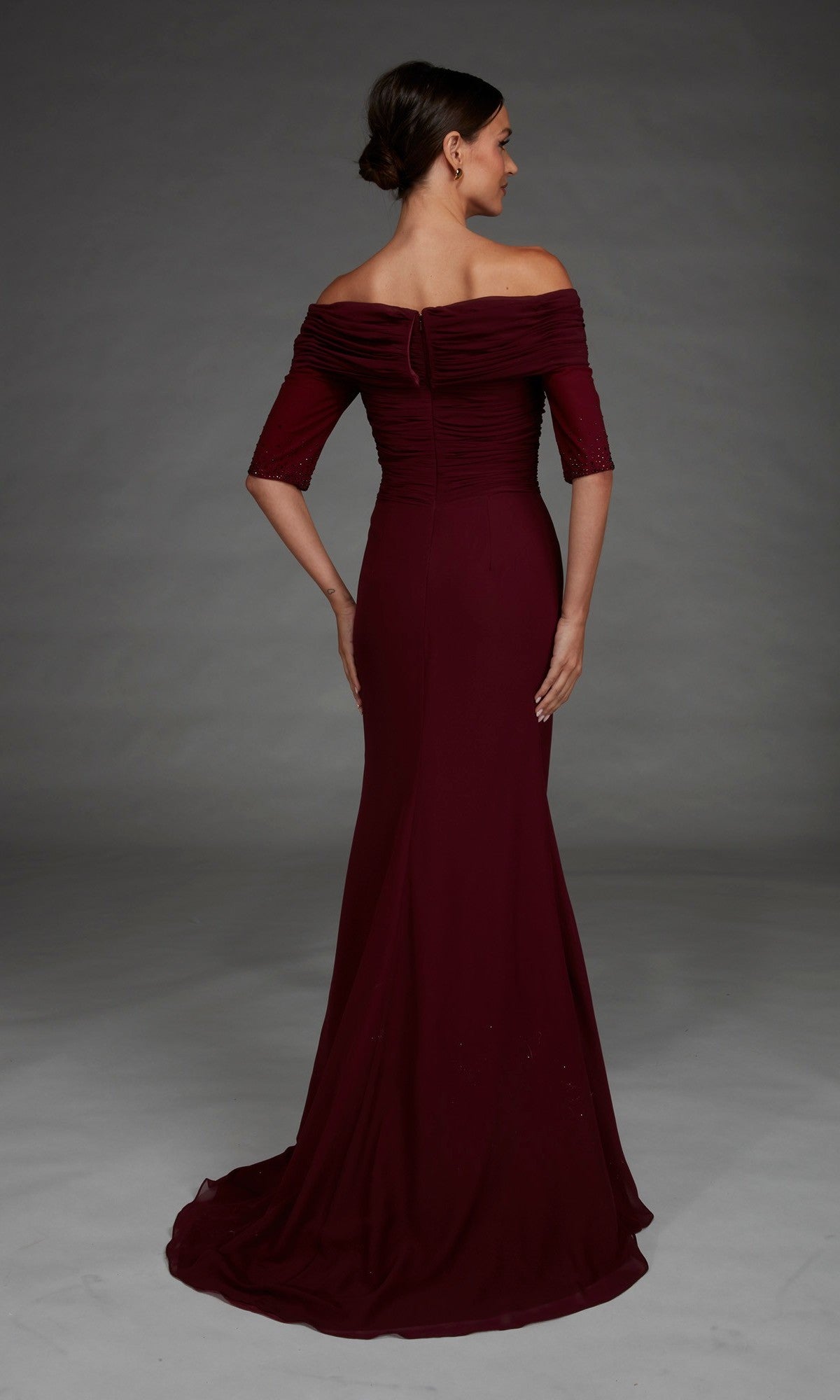 A woman stands with her back to the camera wearing the Long Formal Dress: Alyce 27888, a floor-length evening gown with an off-the-shoulder neckline, fitted sleeves, and an elegant train.