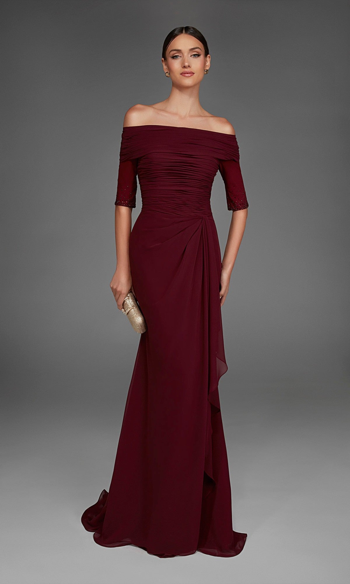 A woman poses against a plain gray background, holding a gold clutch and wearing the Long Formal Dress: Alyce 27888—a floor-length burgundy gown that showcases an elegant off-the-shoulder neckline with short sleeves.