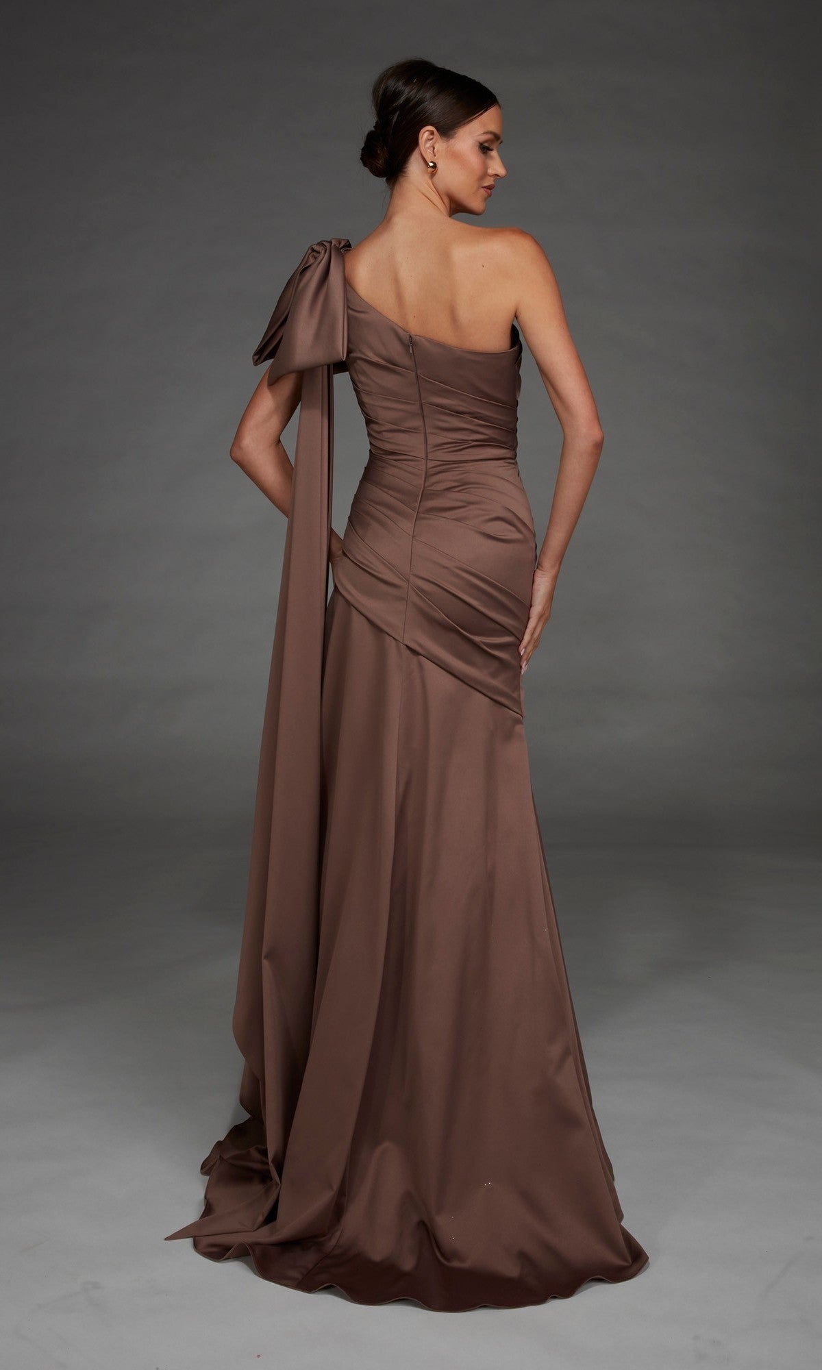 A woman models the Alyce 27891 Long Formal Dress—a floor-length, brown, one-shoulder satin gown featuring an oversized bow and sash detail—as she stands against a plain gray background facing away.