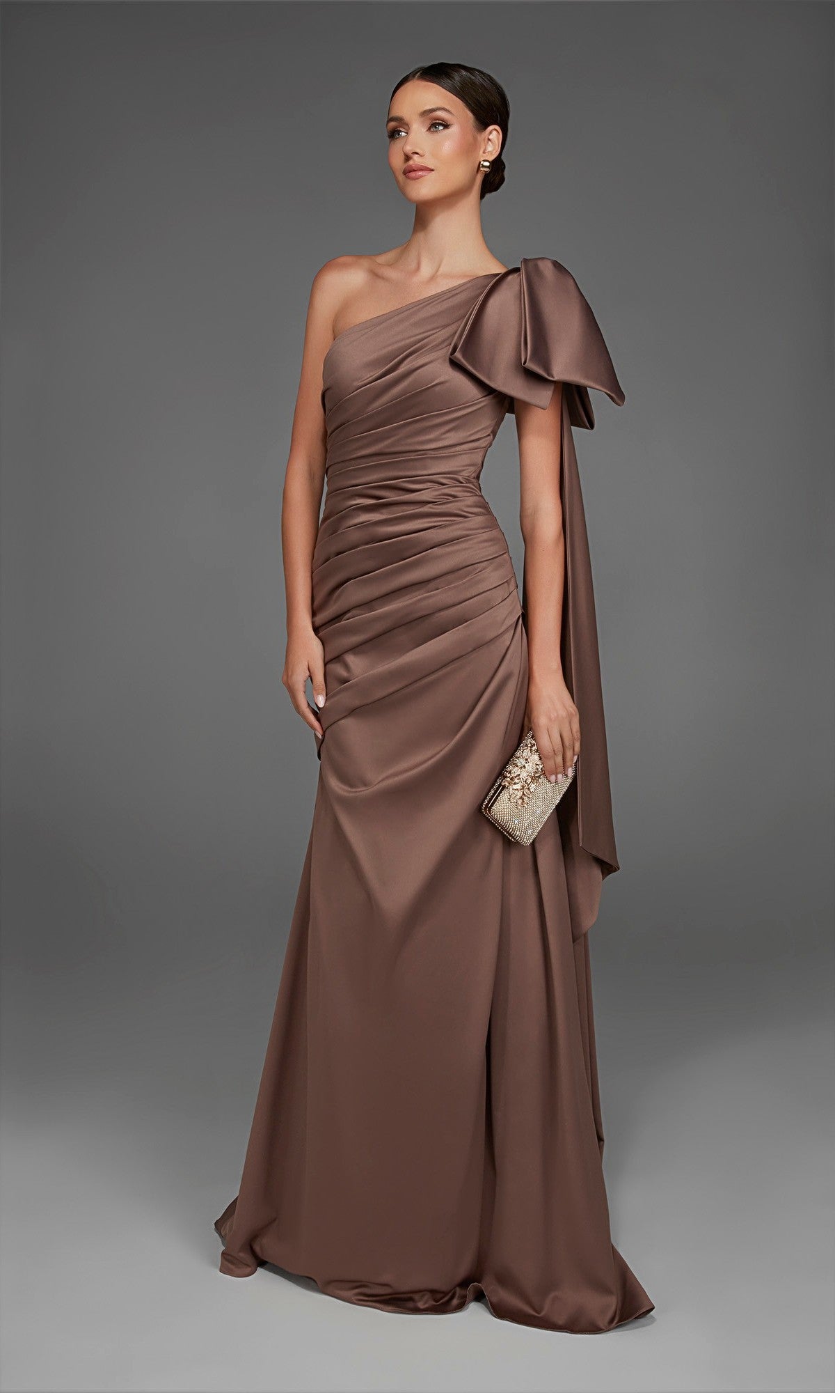A woman models the Long Formal Dress: Alyce 27891—a brown, one-shoulder satin evening gown featuring a dramatic oversized bow on the shoulder—while holding a small clutch against a plain gray background.
