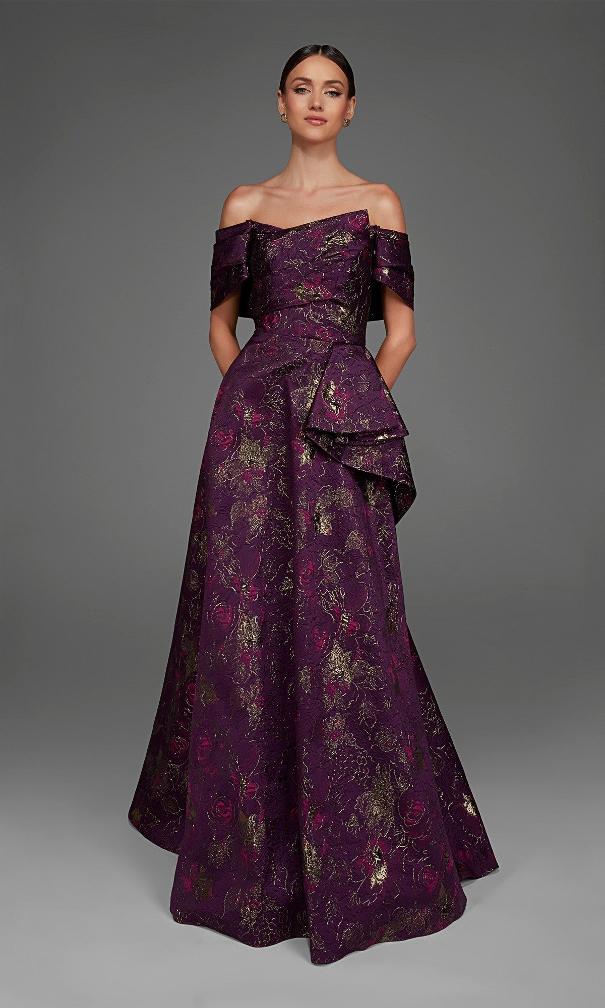 A woman wearing the Long Formal Dress: Alyce 27893 in a rich eggplant hue, featuring elegant off-the-shoulder straps and a luxurious purple and gold floral jacquard pattern, stands against a plain gray background.