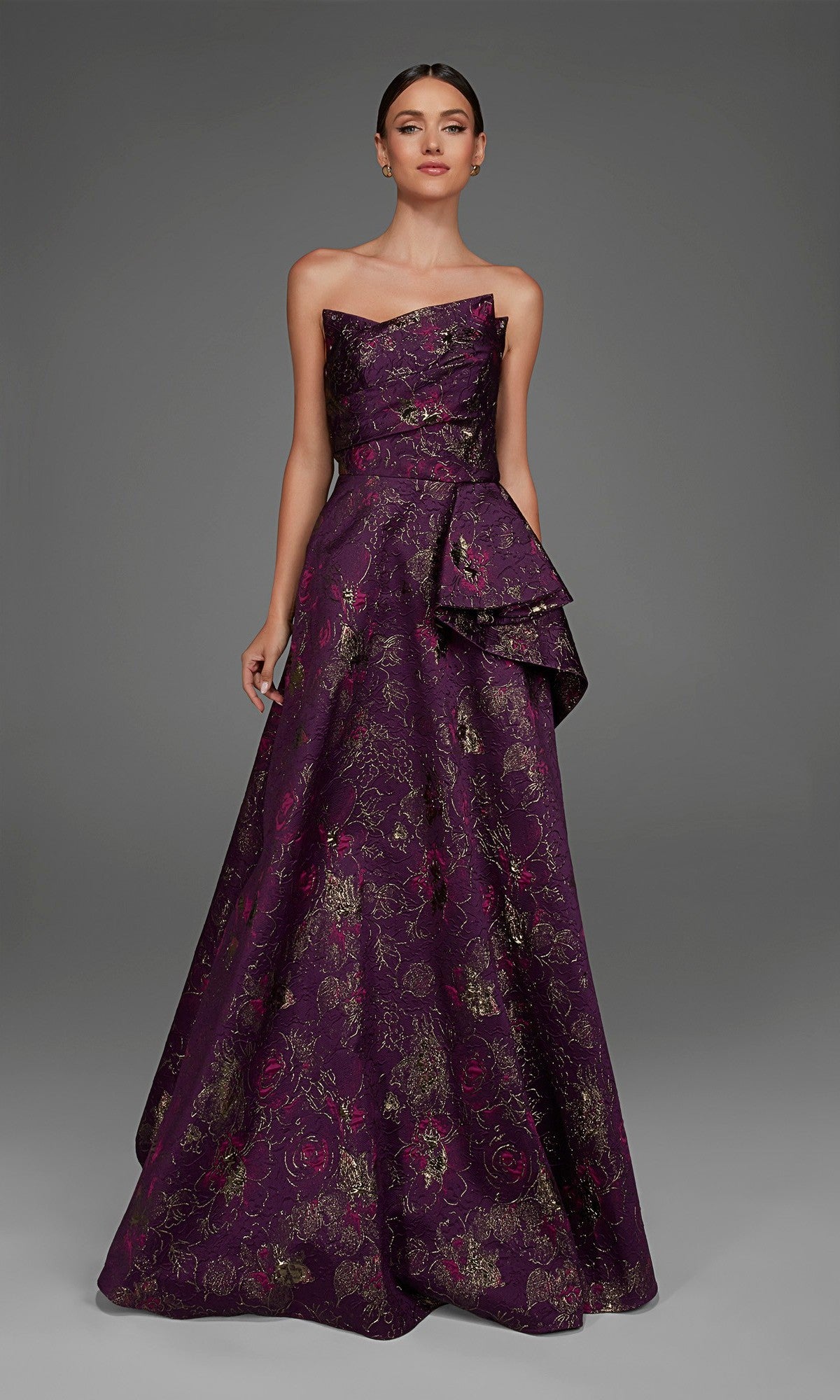 A woman wearing the Alyce 27893 long formal dress, featuring a stunning floor-length silhouette in purple with delicate floral jacquard patterns and a side bow, stands against a plain gray background.