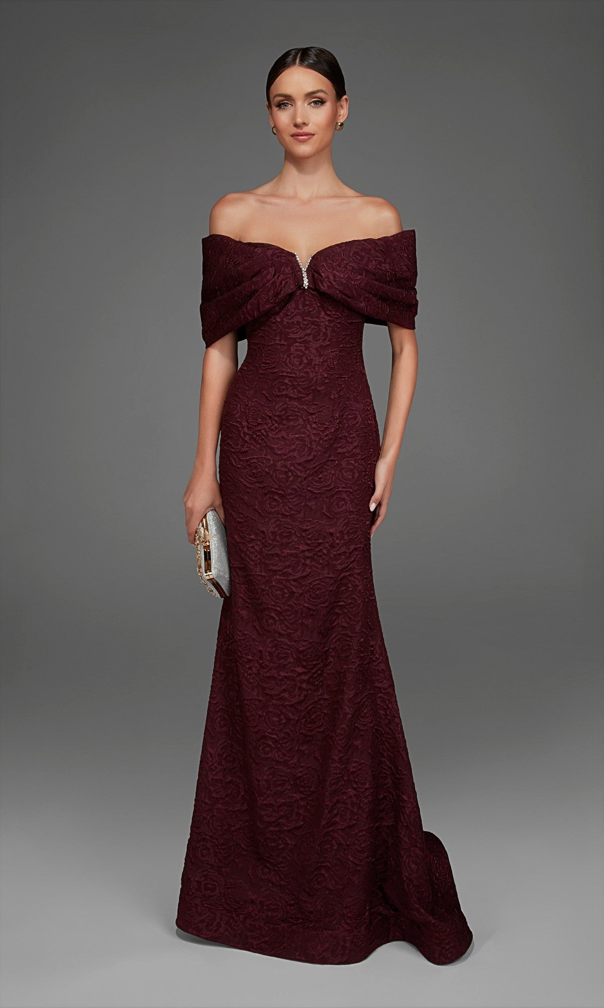 A woman wearing the Long Formal Dress: Alyce 27894 in Black Cherry, featuring an off-the-shoulder, floor-length mermaid silhouette with textured rose-patterned brocade, holds a small metallic clutch and stands against a plain gray background.