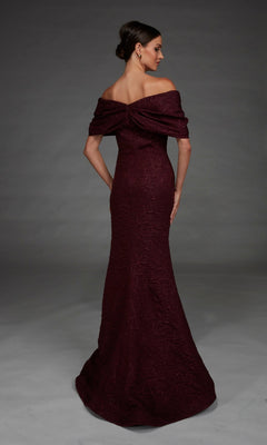 A woman stands with her back to the camera, modeling the Alyce 27894 long formal dress—an off-the-shoulder, floor-length maroon gown featuring a rose-patterned brocade and an elegant mermaid silhouette, set against a plain gray background.