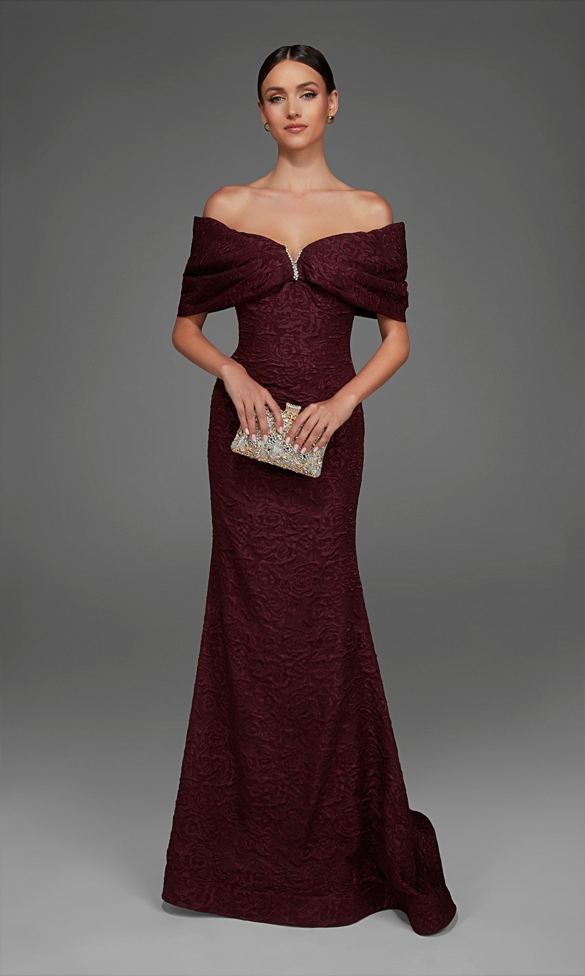 A woman wearing the Alyce 27894 long formal dress—an off-the-shoulder, dark red gown featuring textured fabric and a subtle rose-patterned brocade—holds a jeweled clutch while standing against a plain gray background.