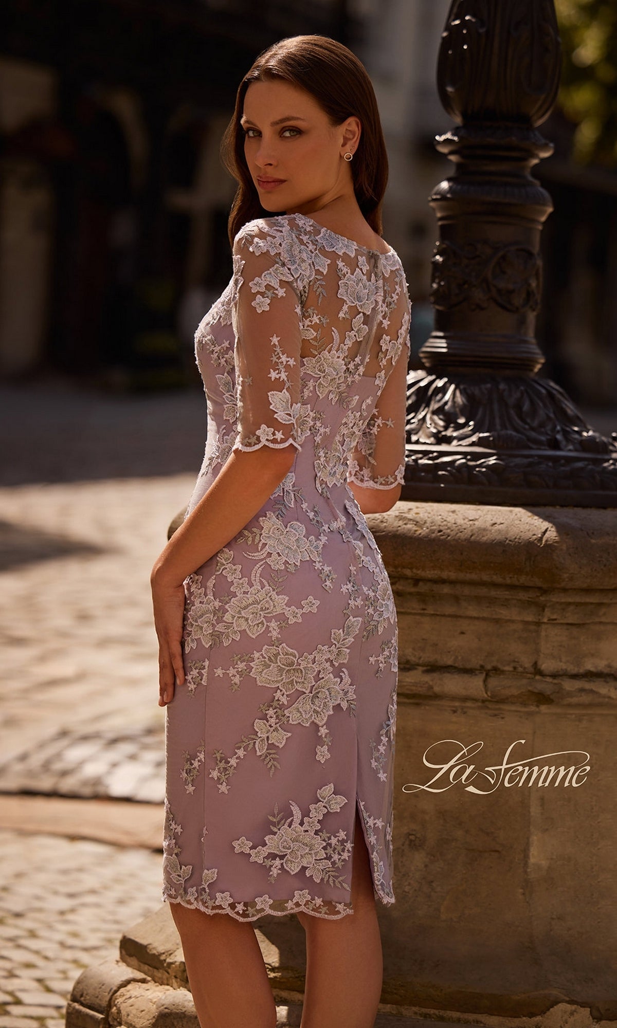 Woman outdoors in Knee Length Wedding Guest Dress: La Femme 27895, lilac floral lace, sheer sleeves, sheath silhouette. Stone pillar, cobblestone ground, “La Femme” text visible.