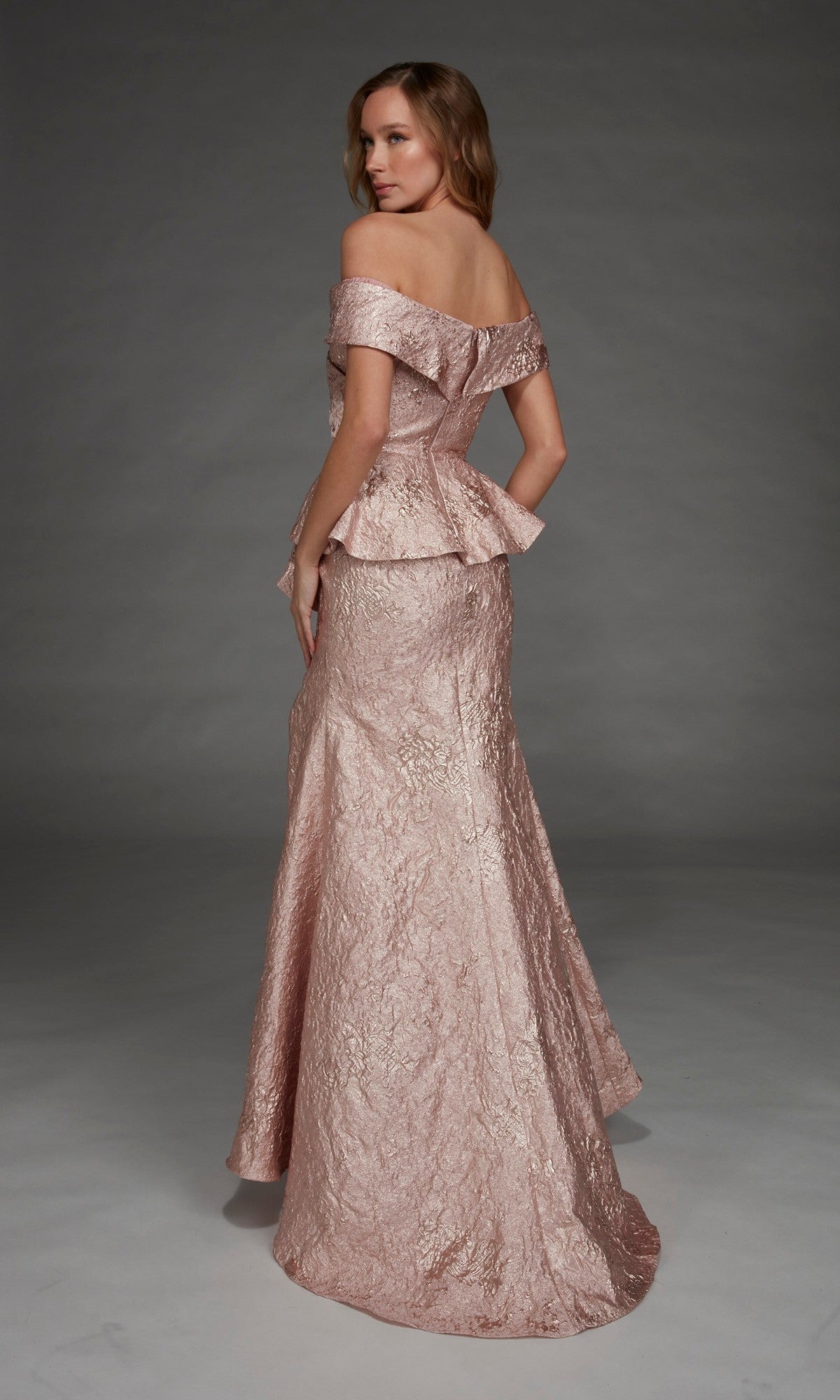 A woman stands against a plain gray background, looking over her shoulder while wearing the Alyce 27899 long formal dress—an off-the-shoulder, textured blush pink gown featuring a peplum waist.