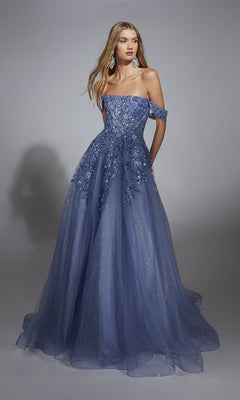 A woman wears the Aerie Couture 278 long prom dress—a blue off-the-shoulder ball gown featuring floral appliqués and lace embroidery on a glitter tulle skirt, standing against a plain gray background.