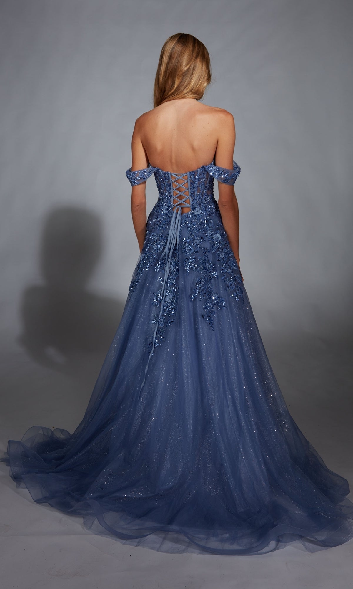 A woman stands with her back to the camera in the Aerie Couture 278 long prom dress, an off-the-shoulder gown with a lace-up back, delicate floral embellishments, and shimmering lace embroidery for an elegant finish.