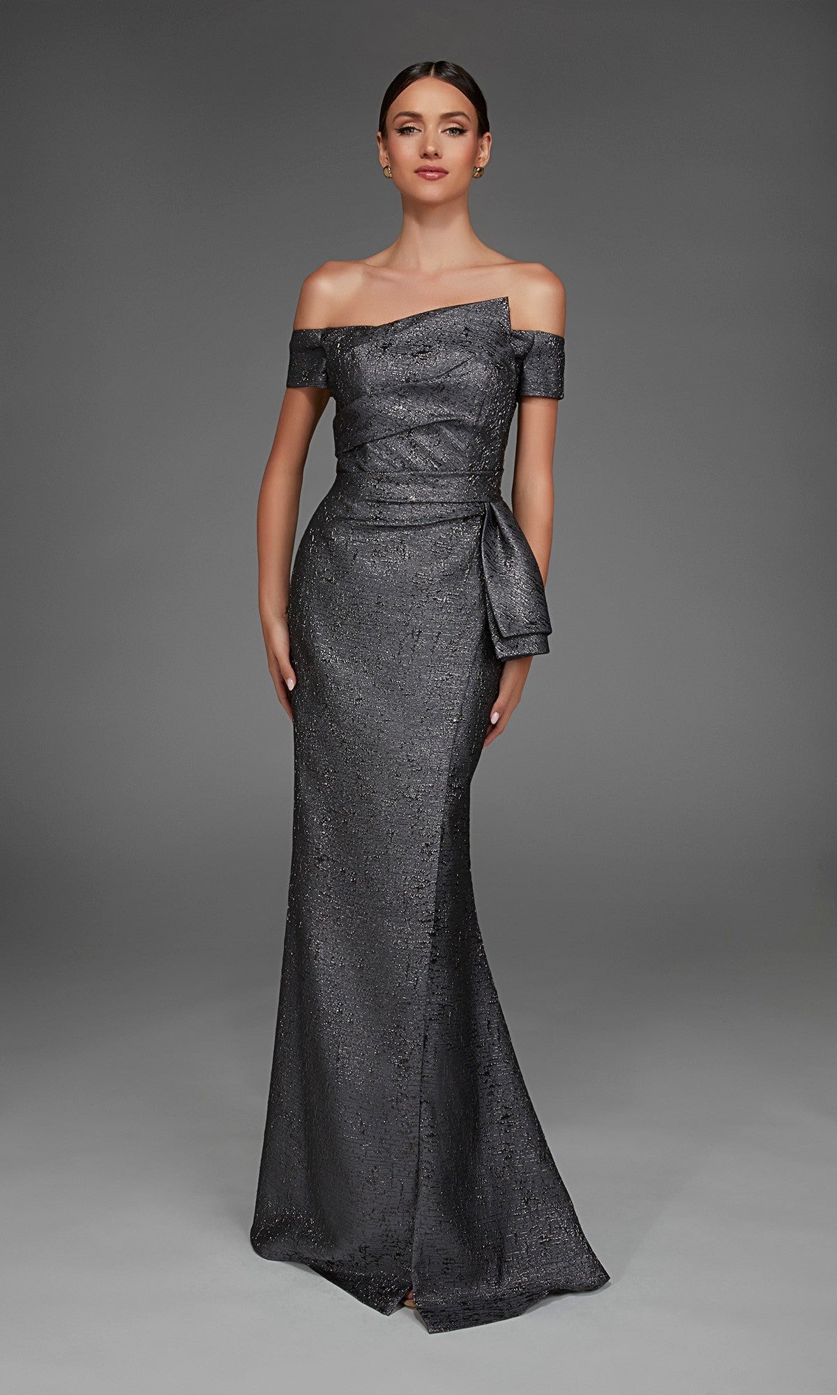 A woman wearing the Alyce 27900 Long Formal Dress in charcoal, featuring an elegant off-the-shoulder metallic jacquard design with a fitted bodice and side bow, stands against a plain gray background.