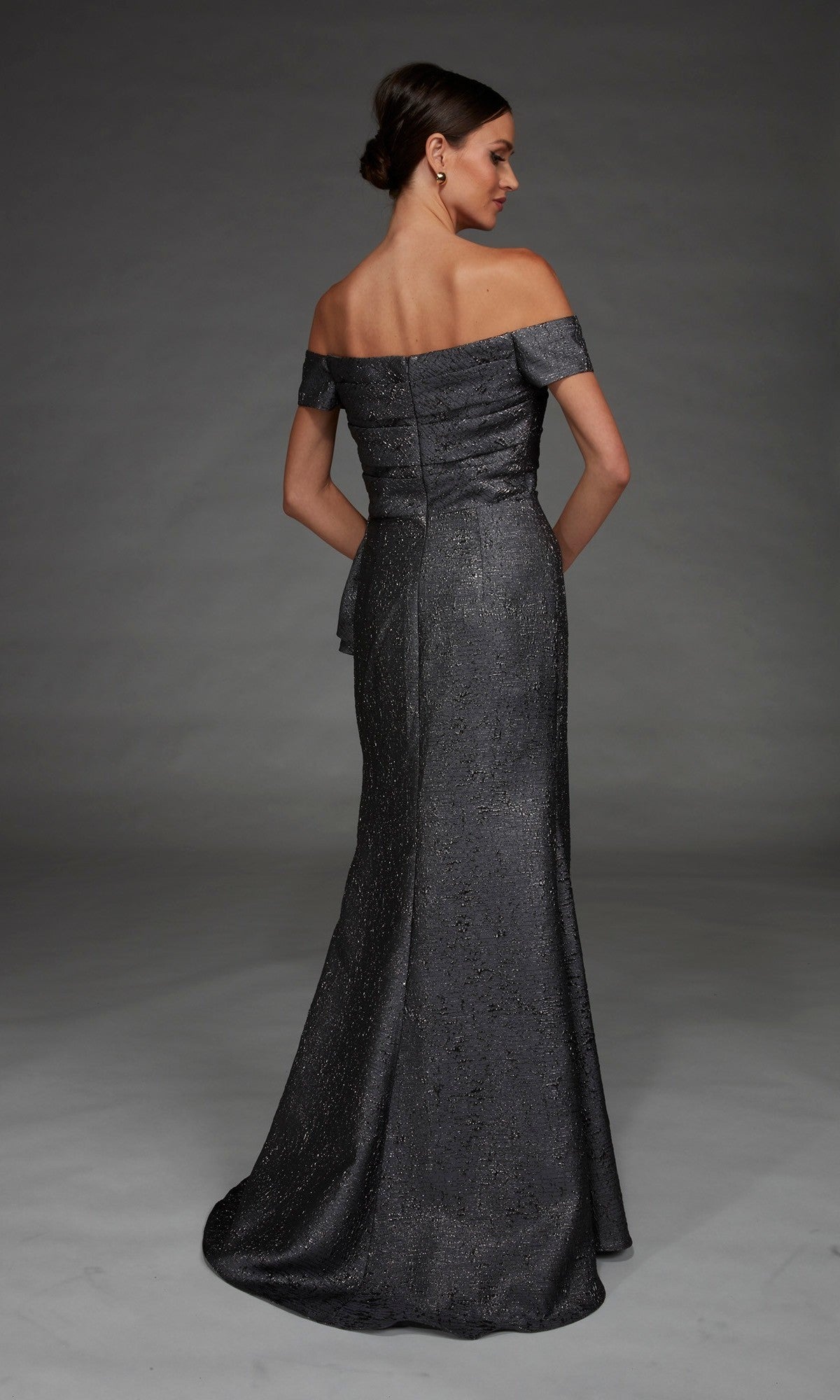 Clad in the Alyce 27900 Long Formal Dress, a woman stands with her back to the camera against a plain gray background, the off-the-shoulder metallic jacquard gown catching the light and shimmering elegantly all the way to the floor.