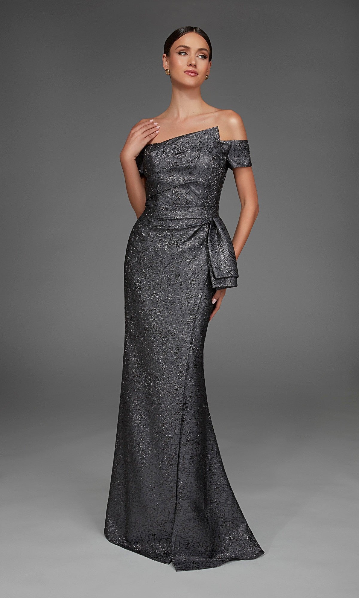 A woman wearing the Alyce 27900 long formal dress—a floor-length, off-the-shoulder metallic jacquard gown with a side bow—stands against a plain gray background.