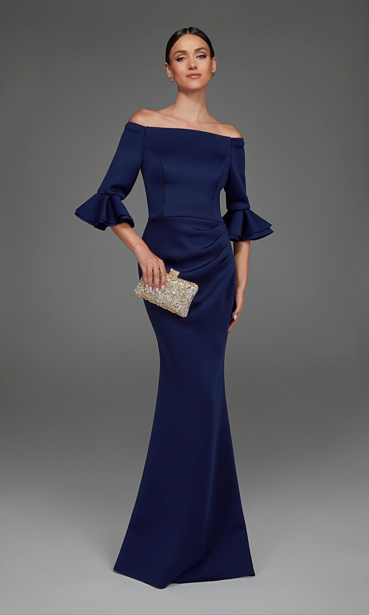 A woman models the Alyce 27905 long formal dress in navy, featuring an off-the-shoulder neckline, ruffled sleeves, and a graceful mermaid skirt. She holds a sparkly clutch while standing against a plain gray background.
