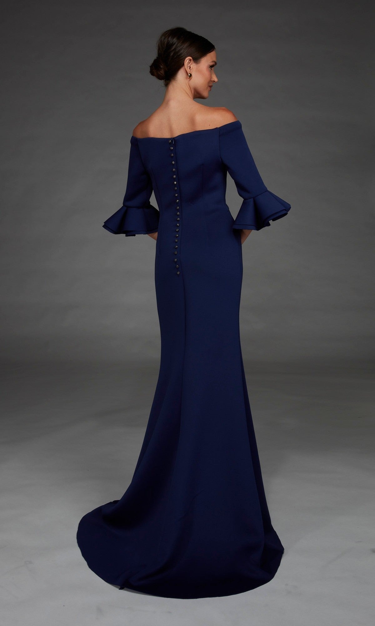 A woman is standing against a gray background, wearing the Alyce 27905—a navy blue, off-the-shoulder long formal dress with ruffled sleeves, a button-up back, and an elegant mermaid skirt.
