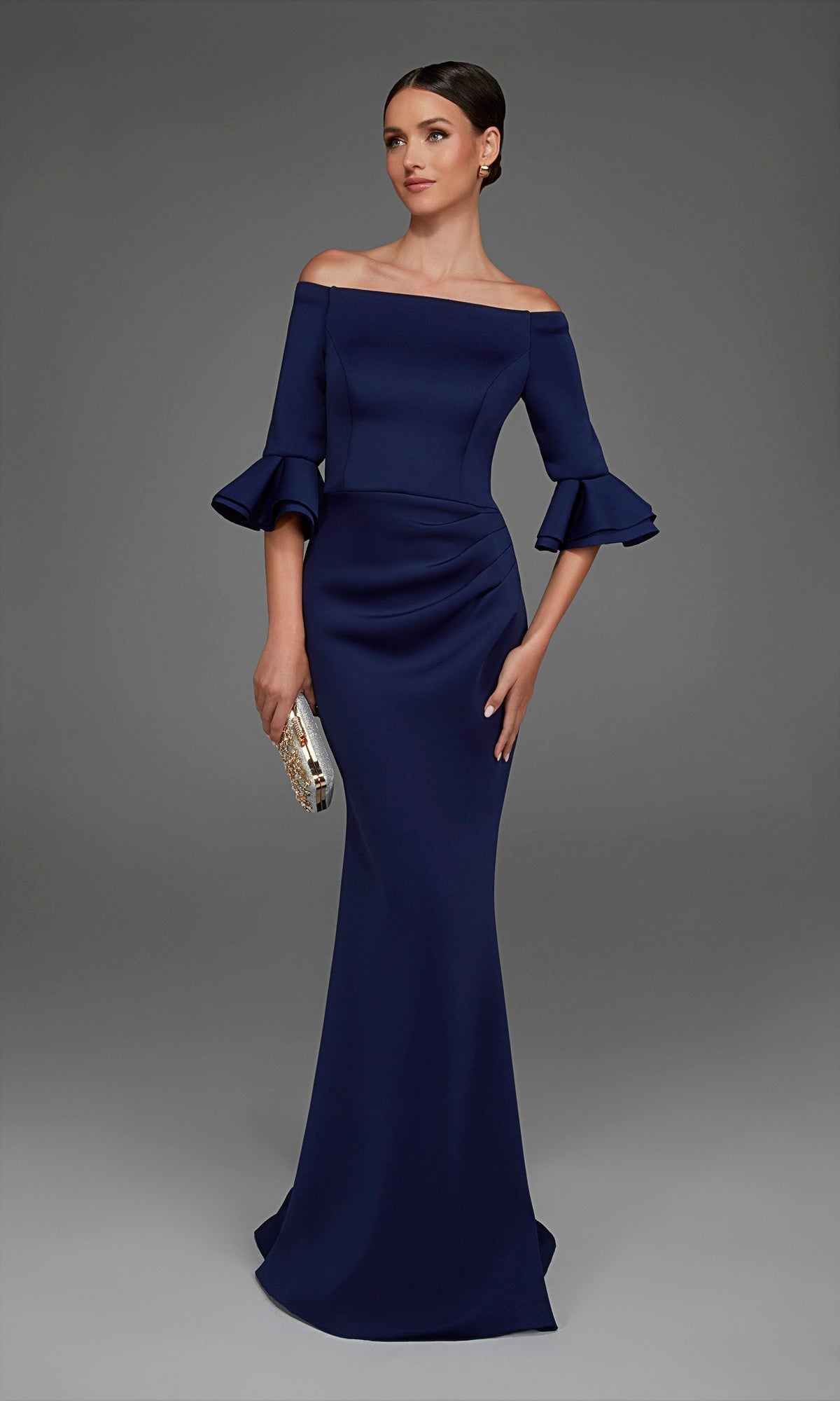 Wearing the Alyce 27905 long formal dress, a woman poses against a plain gray background. The off-the-shoulder navy blue gown features tiered ruffle sleeves and a graceful mermaid skirt, which she pairs with a gold clutch.
