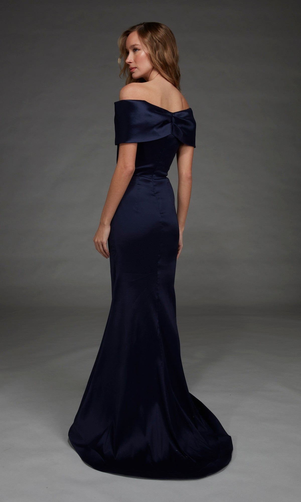 A woman wearing the Alyce 27908 Long Formal Dress— a floor-length, off-the-shoulder gown crafted from stretch mikado with a bow detail on the back—stands in a studio with a plain gray background.