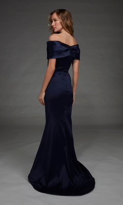 A woman wearing the Alyce 27908 Long Formal Dress— a floor-length, off-the-shoulder gown crafted from stretch mikado with a bow detail on the back—stands in a studio with a plain gray background.