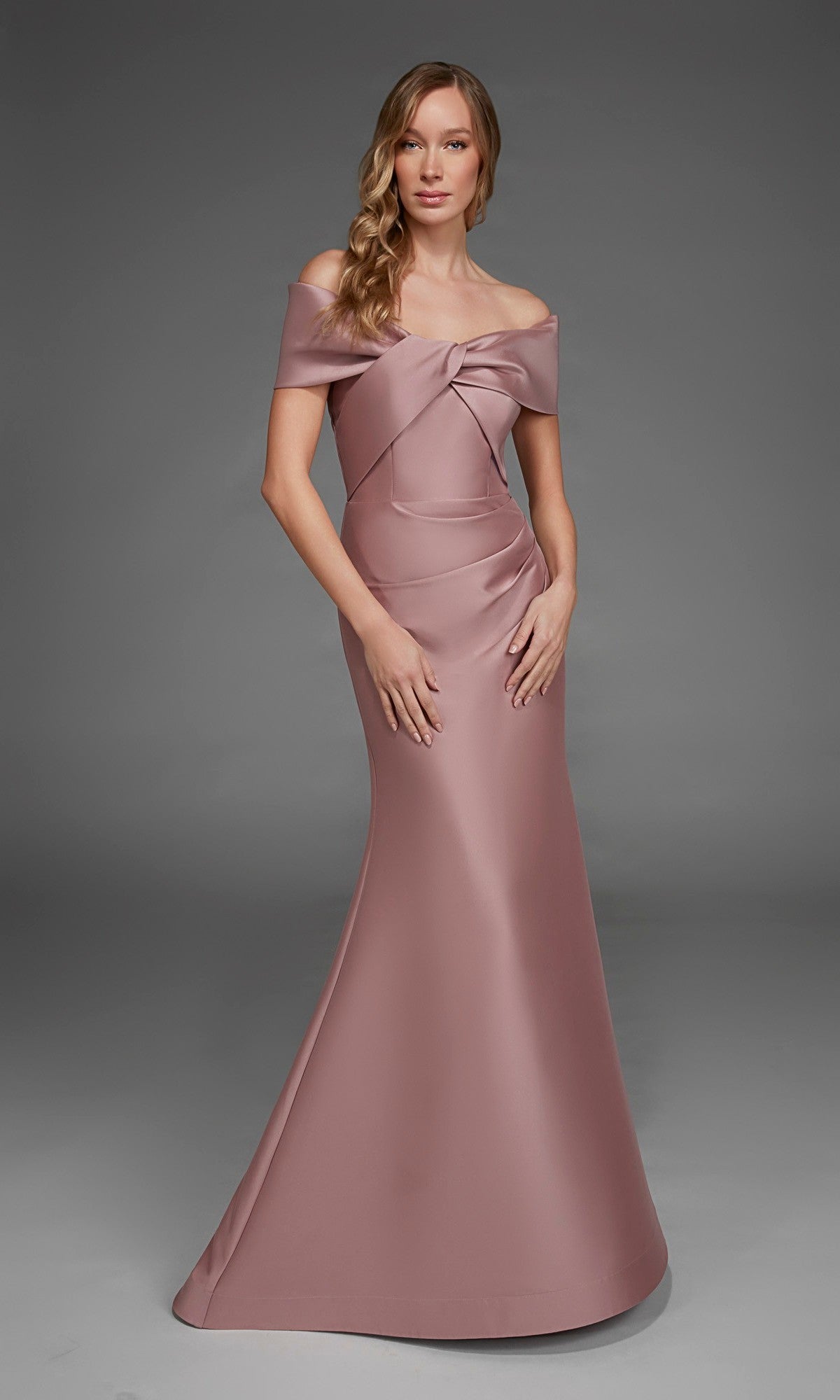 A woman wears the Long Formal Dress: Alyce 27908, a mauve, off-the-shoulder, floor-length gown crafted from luxurious stretch mikado with a large bow detail on the bodice, as she stands against a plain gray background.