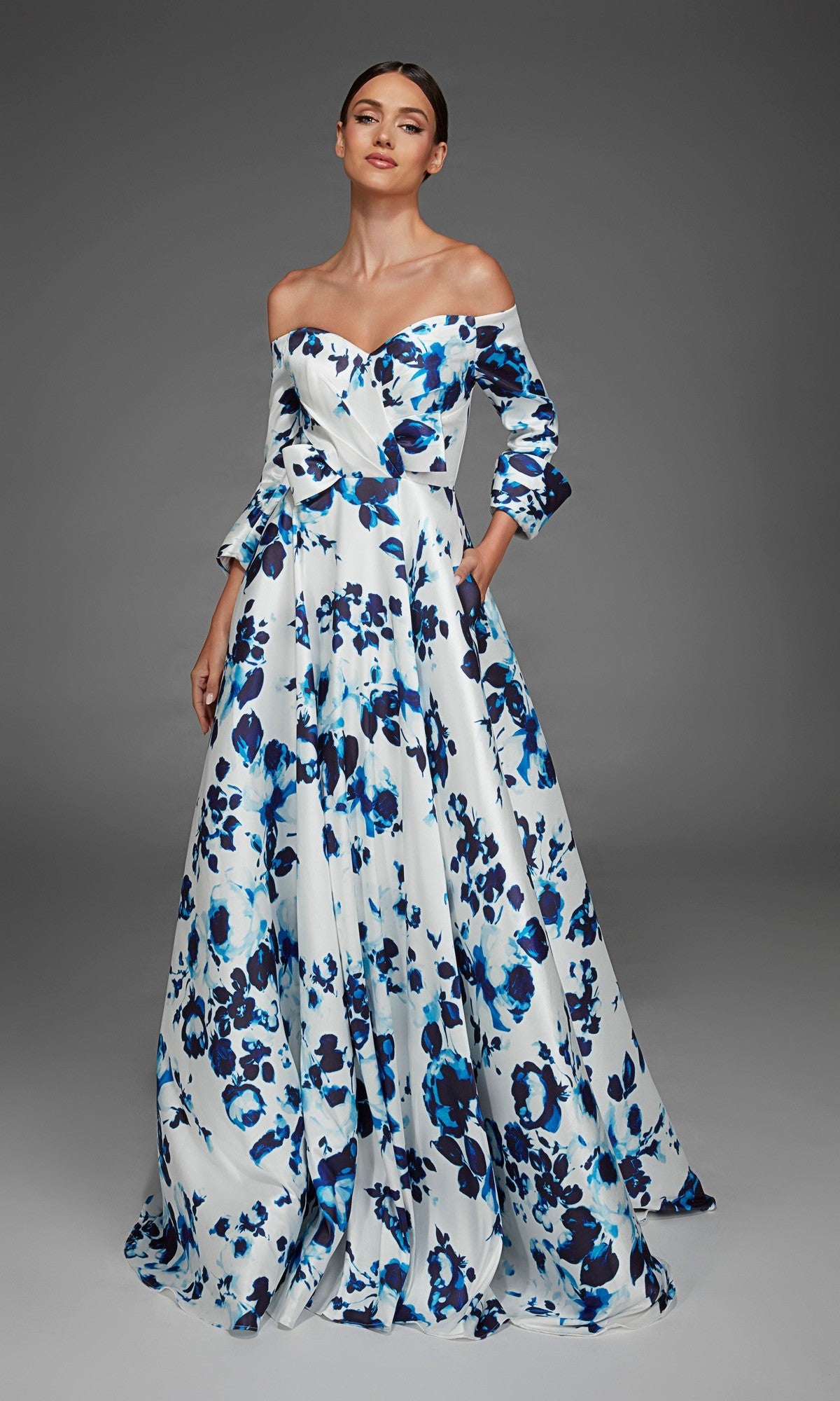 Dressed in the Alyce 27910 long formal gown, the woman exudes elegance in this off-the-shoulder, floor-length creation. The blue and white evening dress features long sleeves and a graceful floral print that skims her silhouette as she stands against a plain gray background.