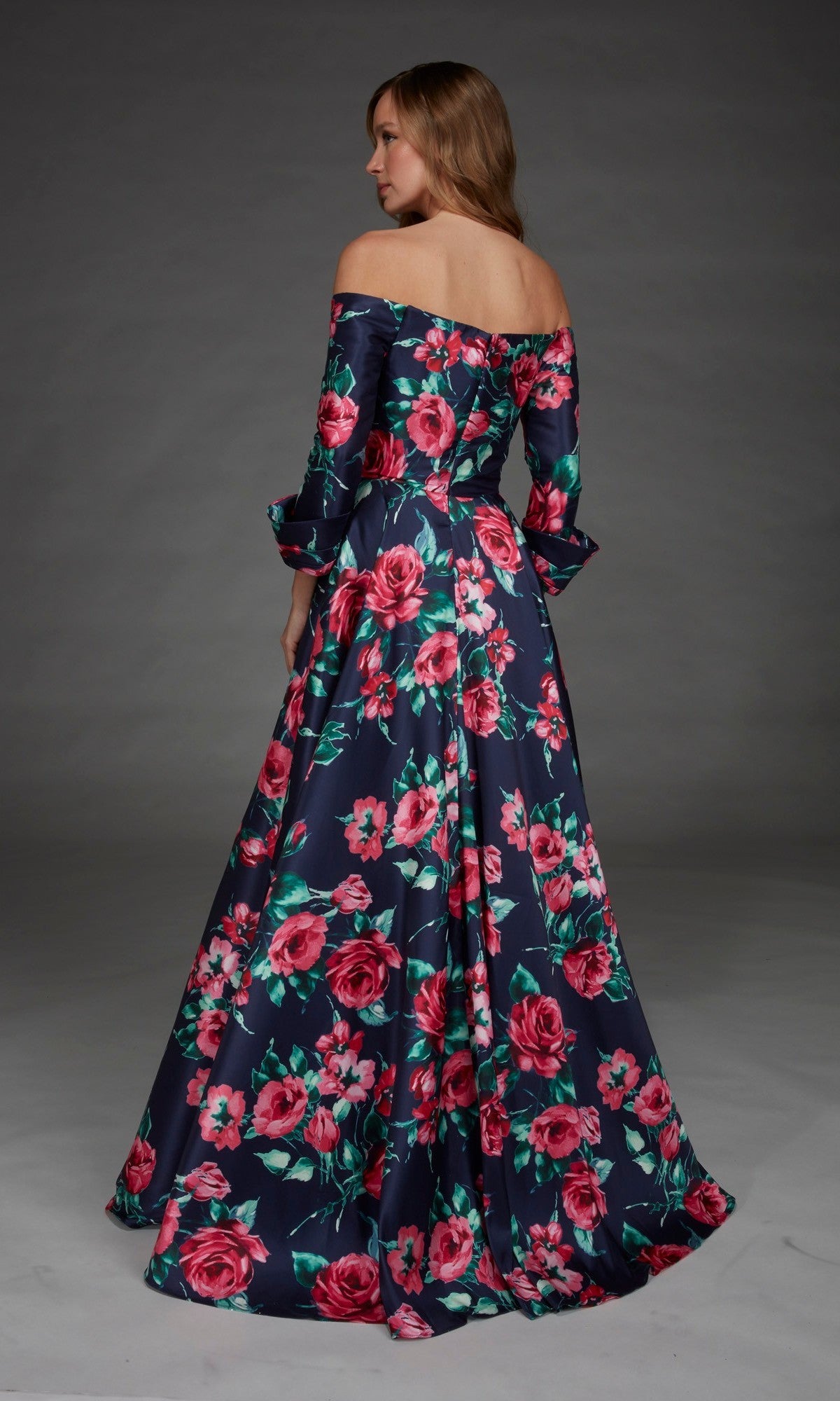 A woman stands facing away from the camera wearing the Alyce 27910 Long Formal Dress, a floor-length navy evening gown featuring an off-the-shoulder design and a pink floral pattern.