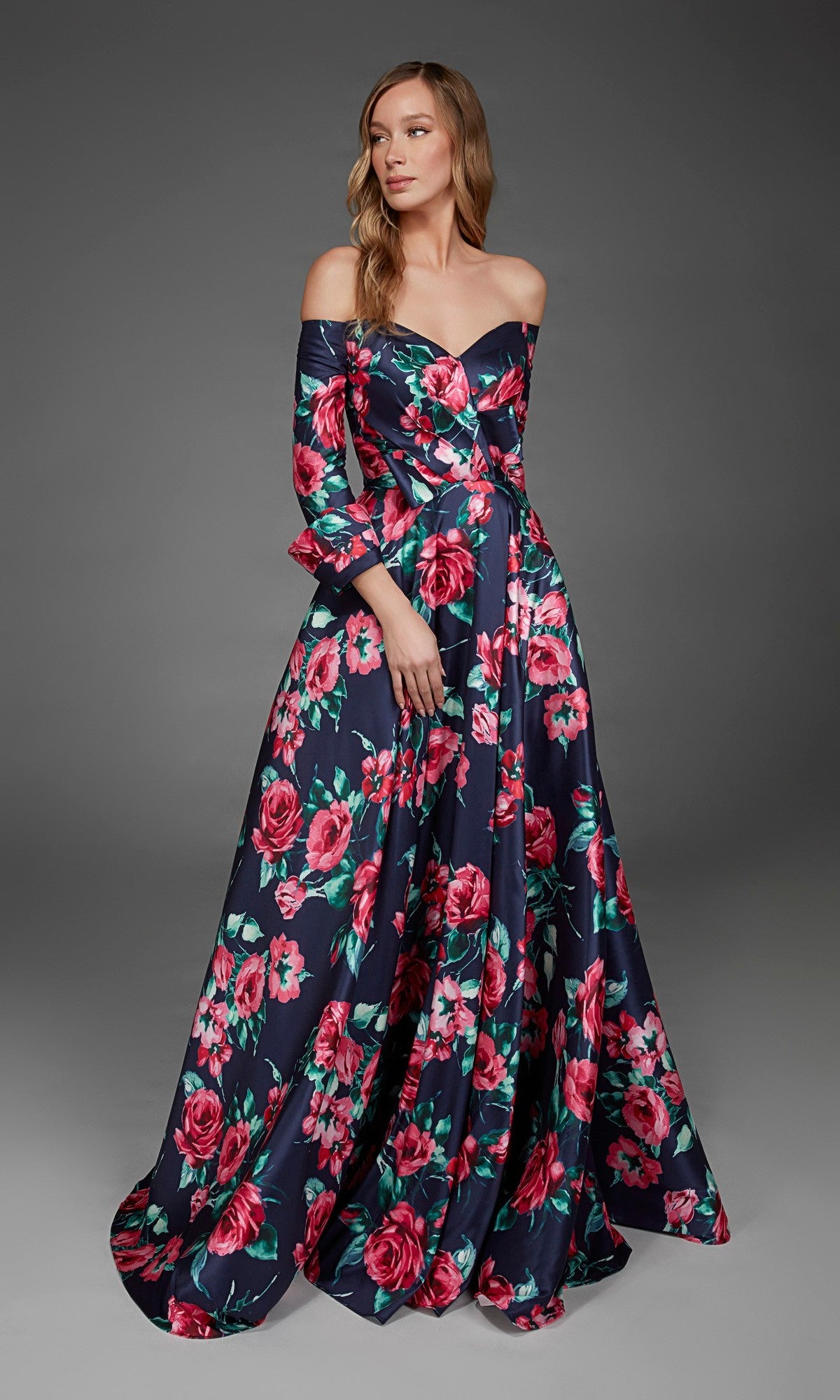 A woman stands against a plain dark background wearing the Long Formal Dress: Alyce 27910—a navy blue off-the-shoulder gown featuring a bold pink floral print and an elegant a-line skirt.