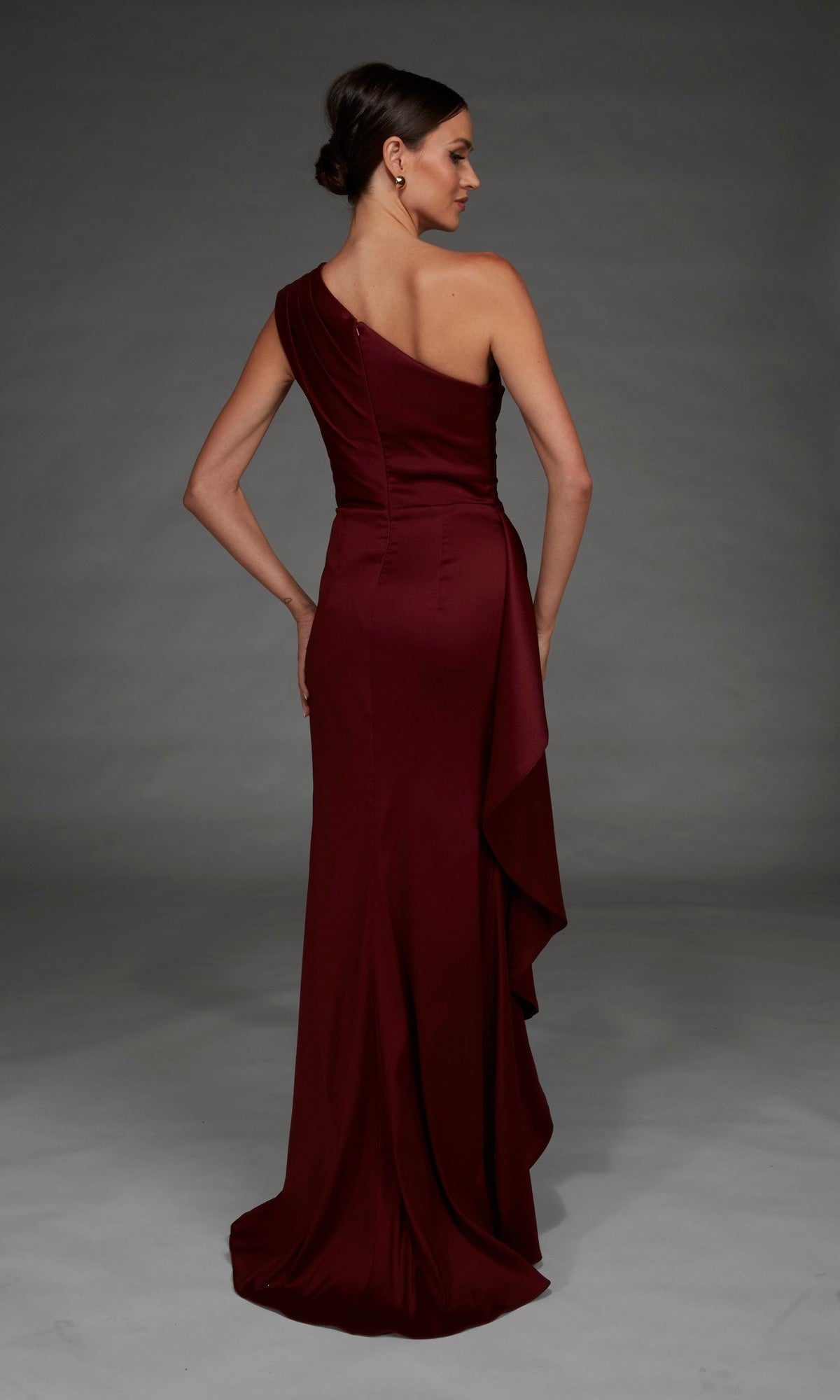 A woman wearing the Alyce 27913 long formal dress, a satin, floor-length, one-shoulder maroon gown, stands facing away with her hair styled in a low bun against a plain gray background.