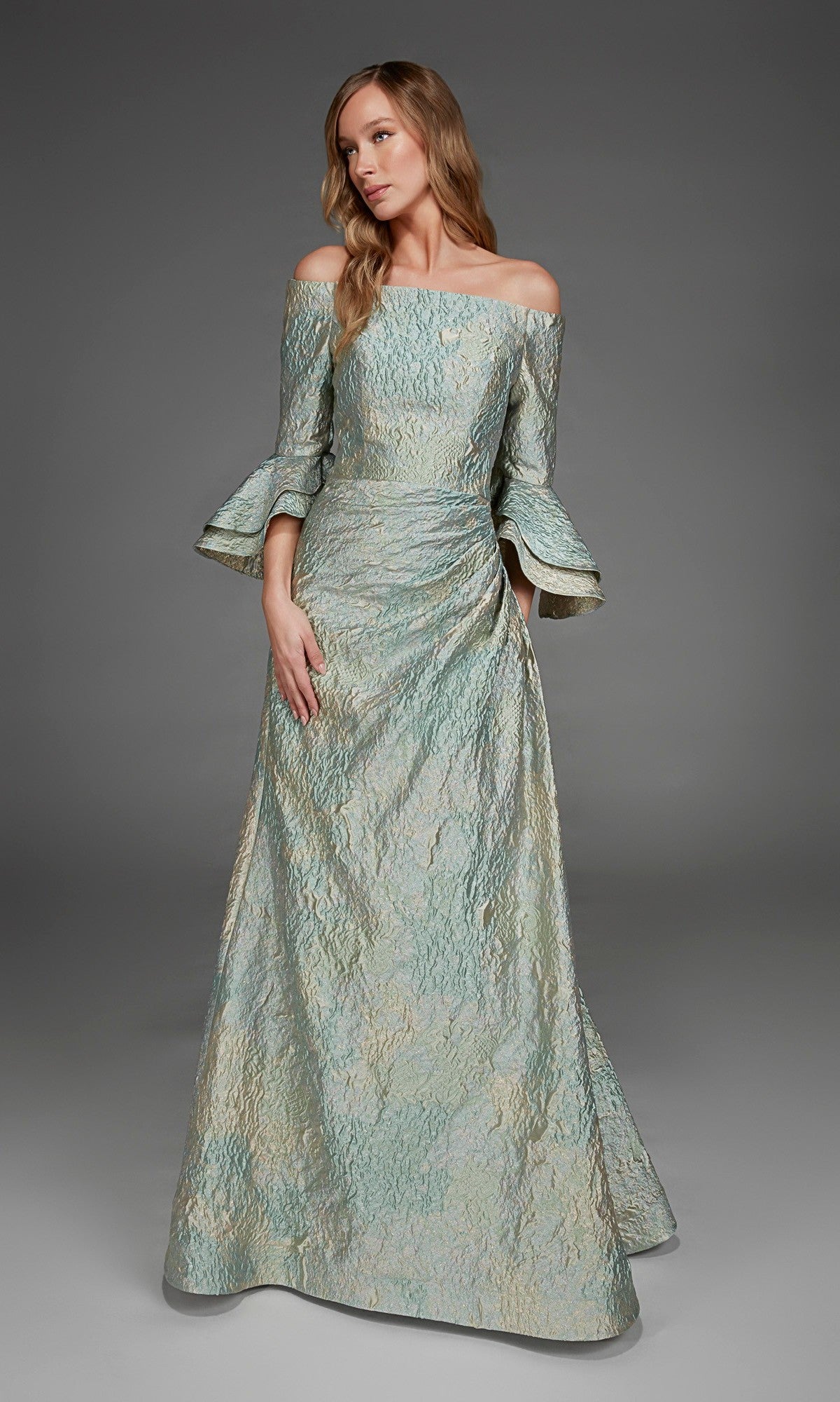 Wearing the Alyce 27914 long formal dress in celadon, a woman stands against a plain gray background. The textured, off-the-shoulder gown features flared sleeves and an A-line silhouette, while its metallic jacquard fabric adds subtle shimmer to the sophisticated look.