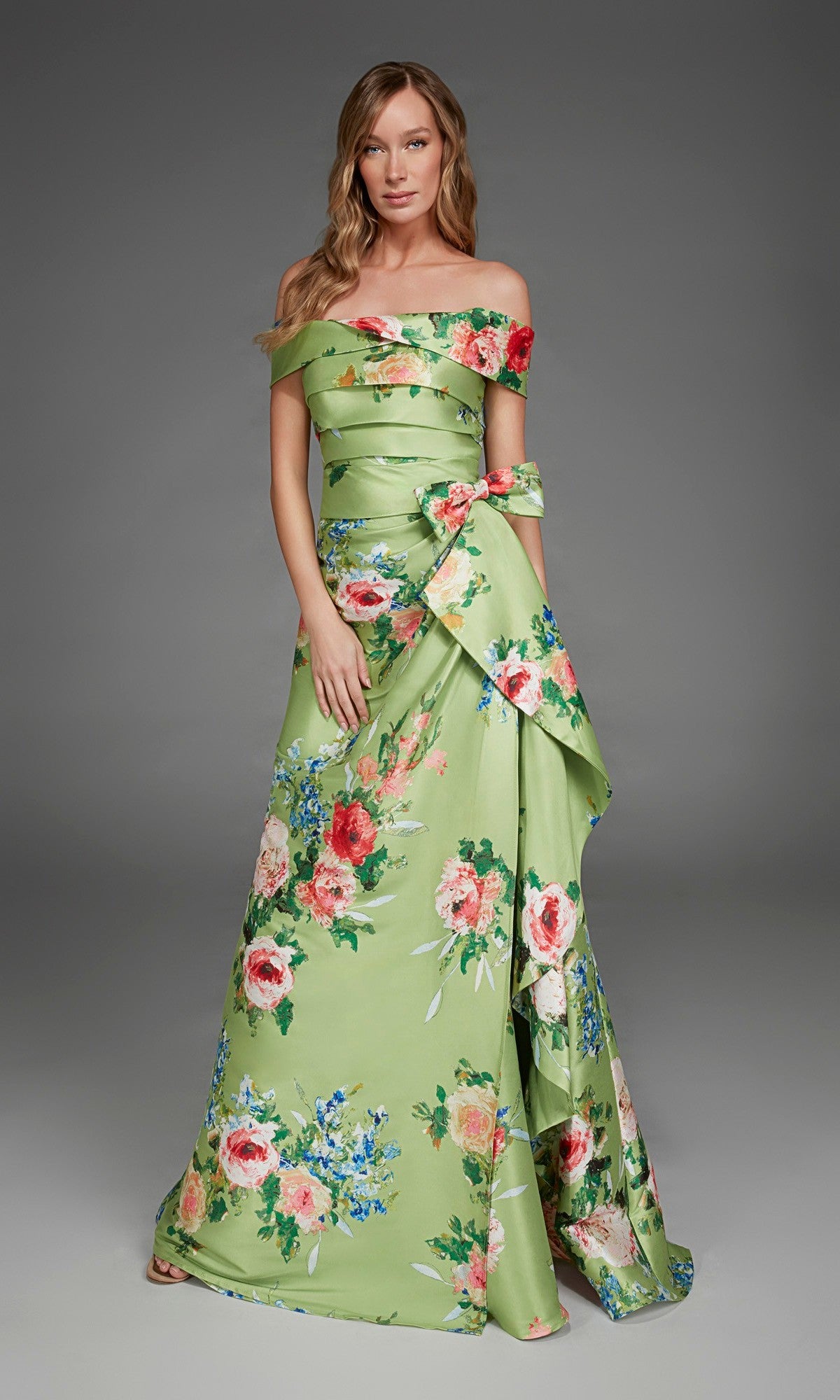 A woman wearing the Alyce 27915 long formal dress in peridot green, featuring an off-the-shoulder design and embellished with red and blue floral accents, stands against a plain gray background.