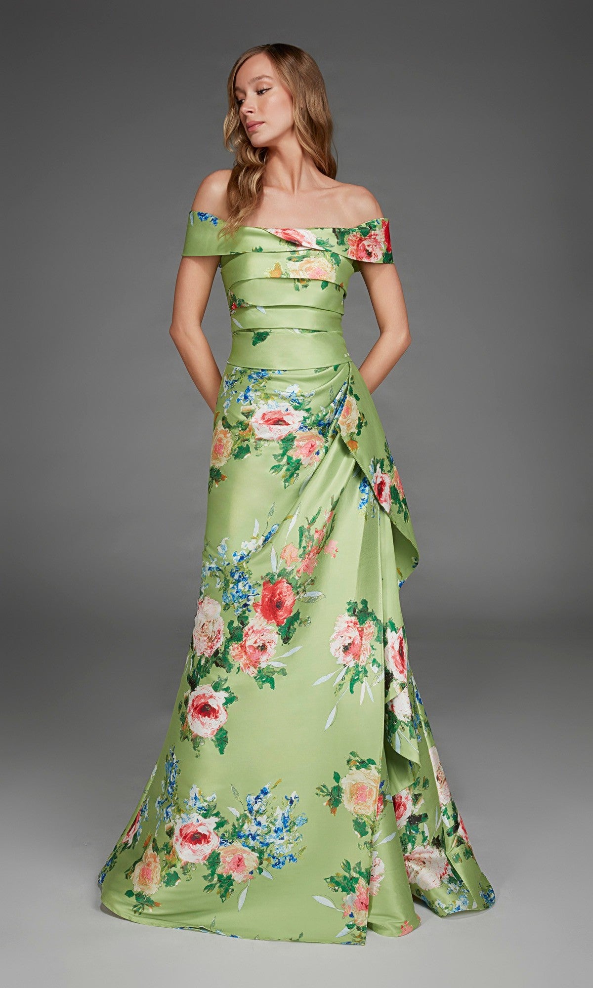 A woman stands wearing the Long Formal Dress: Alyce 27915, an off-the-shoulder, floor-length green satin gown adorned with pink and blue floral prints. She poses with her hands in the dress pockets against a plain gray background.
