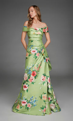A woman stands wearing the Long Formal Dress: Alyce 27915, an off-the-shoulder, floor-length green satin gown adorned with pink and blue floral prints. She poses with her hands in the dress pockets against a plain gray background.