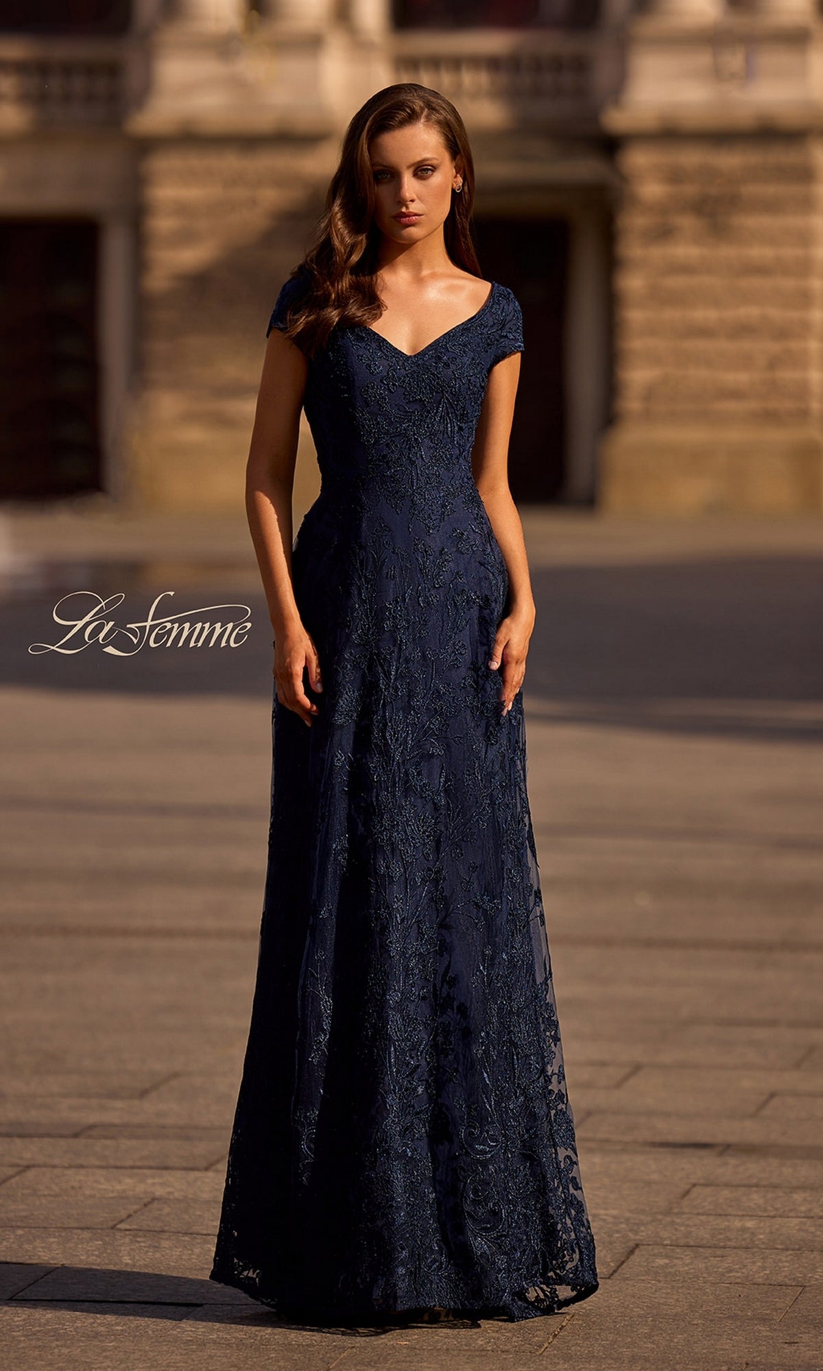 A woman stands outdoors on a stone path in front of a building, wearing the Long Formal Dress: La Femme 27915 in navy with subtle floral patterns.