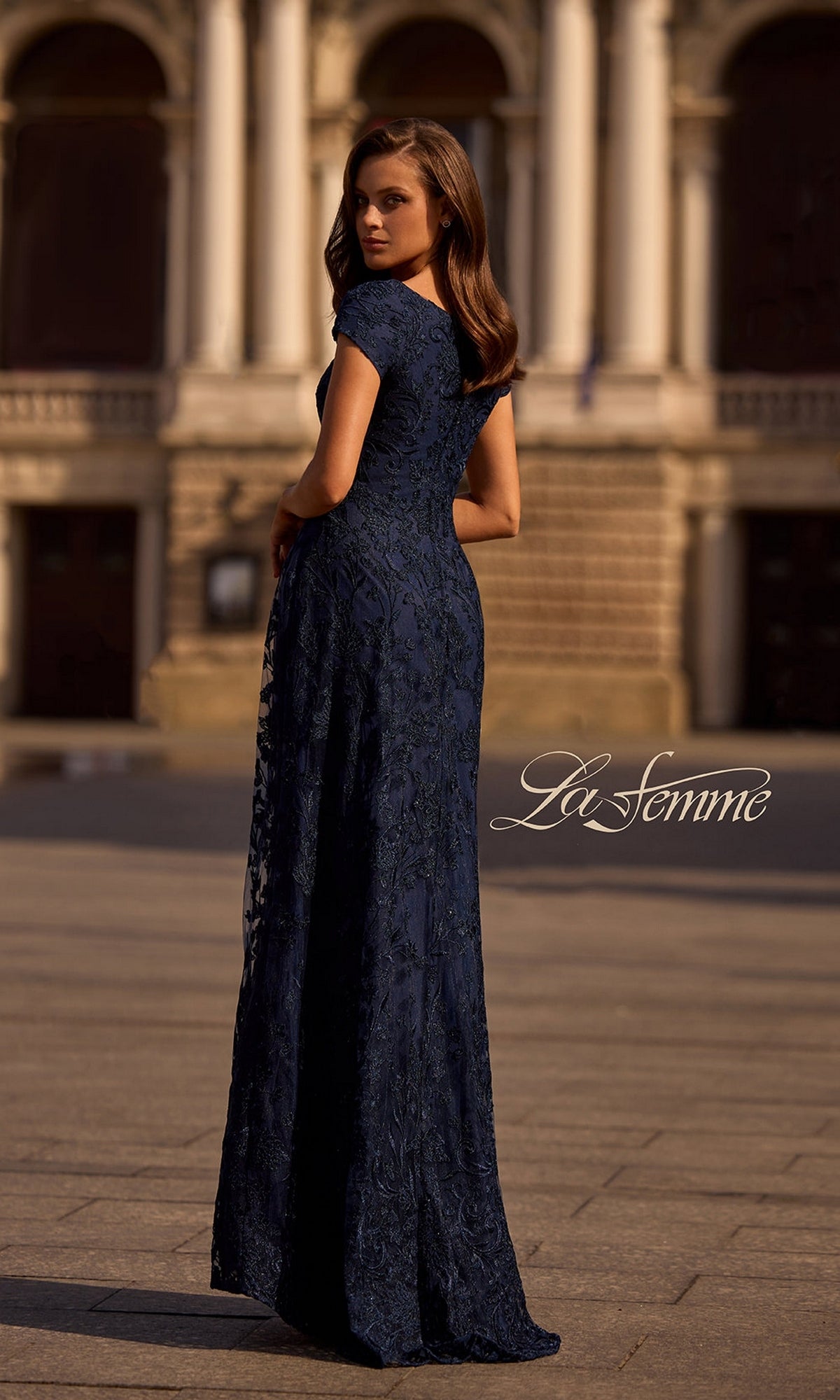 A woman in a navy blue Long Formal Dress: La Femme 27915 poses outdoors by a stone building, glancing back. The “La Femme” logo appears at the lower right.