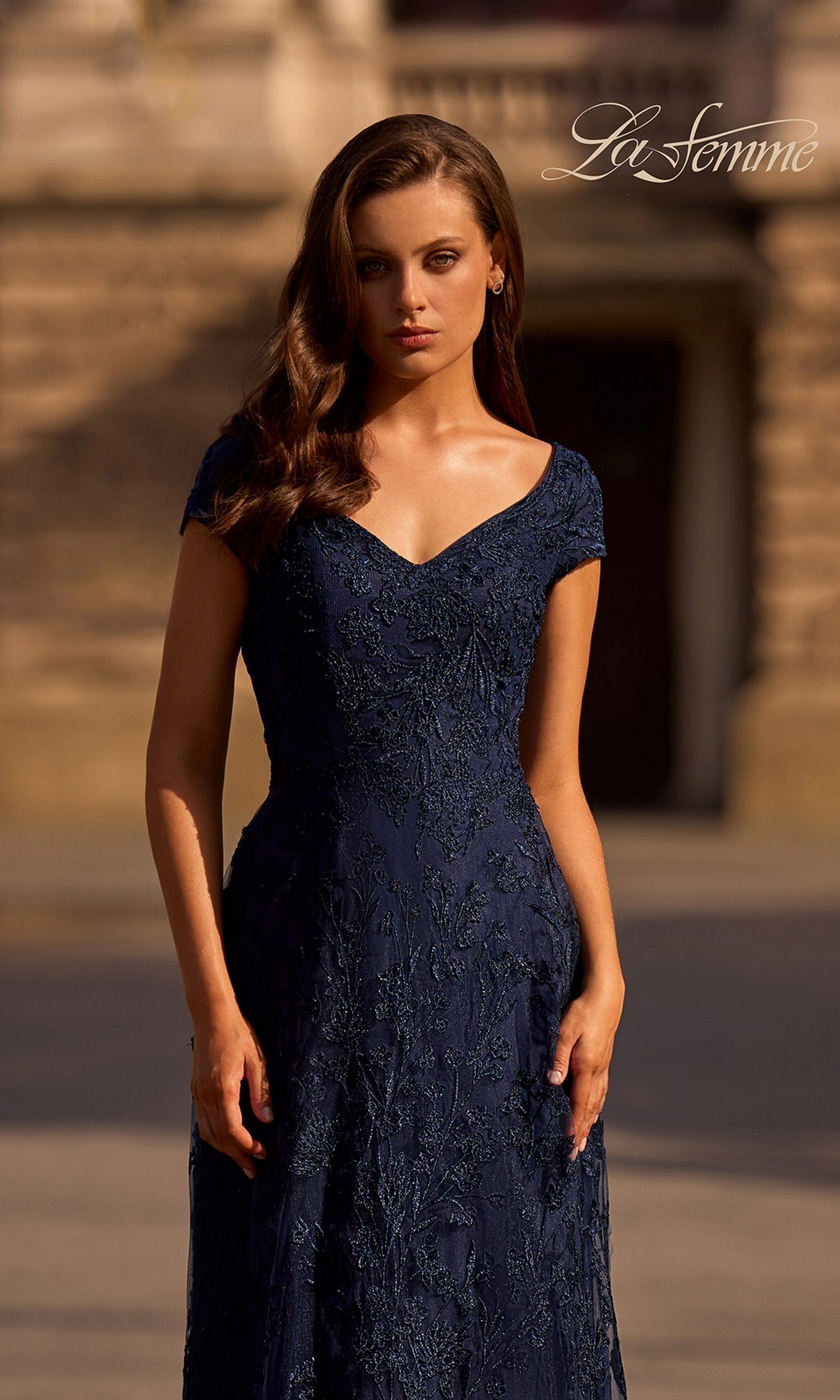 A woman stands outdoors in a navy blue Long Formal Dress: La Femme 27915, with a blurred stone building behind and the “La Femme” logo in the upper right corner.